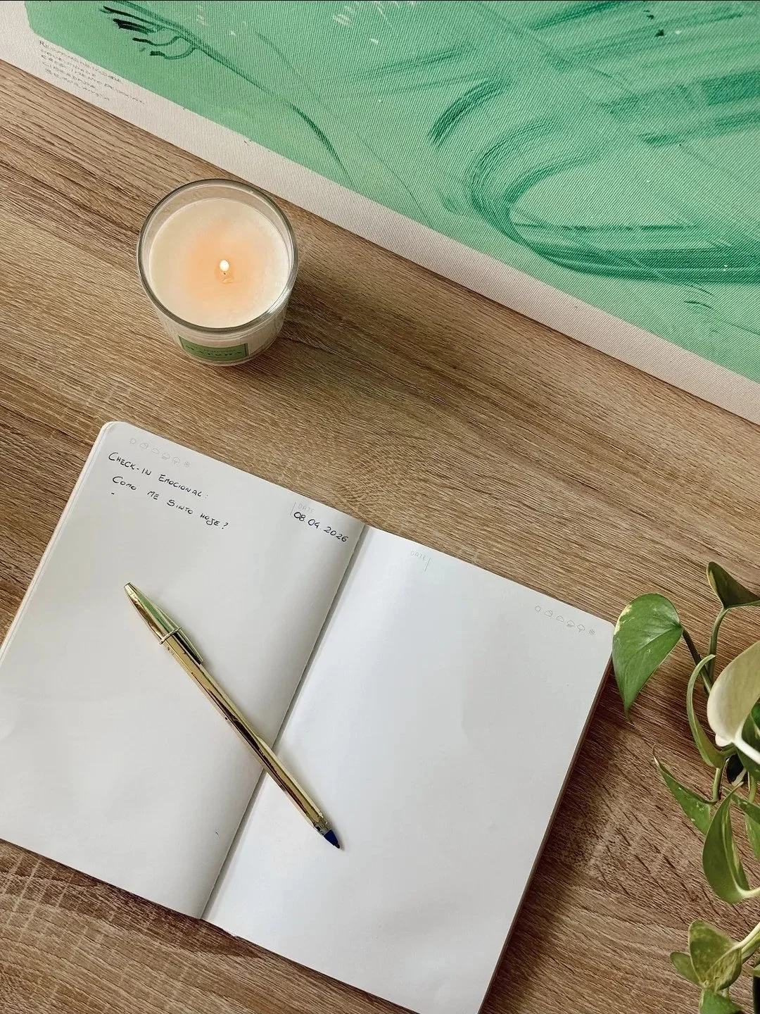 Caderno aberto com uma caneta em cima, uma vela acesa e uma planta de folhas verdes na mesa de madeira, com uma obra de arte verde ao fundo.