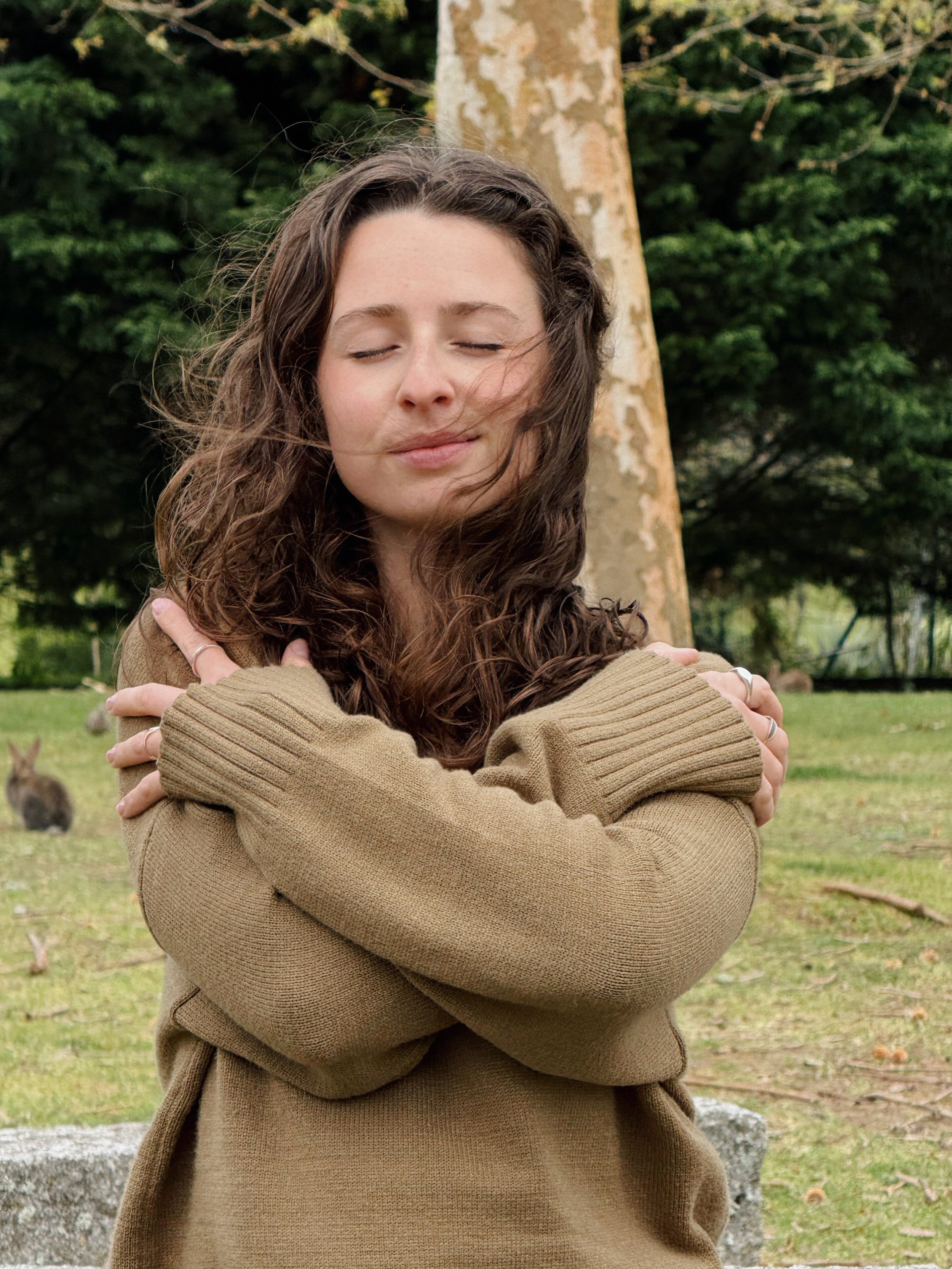 Mulher com cabelo castanho, cacheado e olhos fechados, abraçando-se, usando suéter cinza, ao ar livre em um parque com árvores e grama.