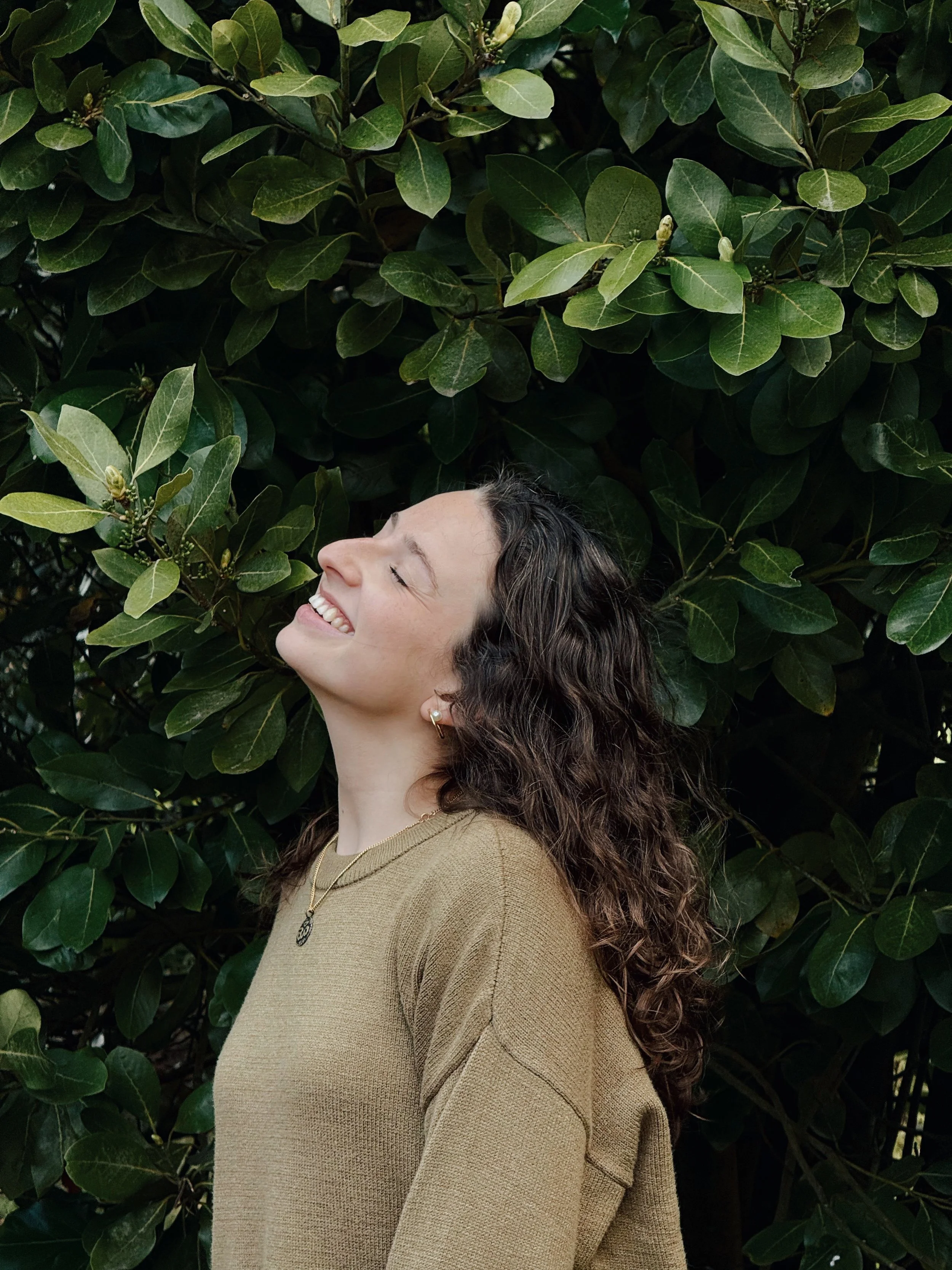 Mulher sorrindo com olhos fechados, cabelo cacheado, usando uma blusa bege, acessórios dourados, em frente a uma grande folhagem verde.