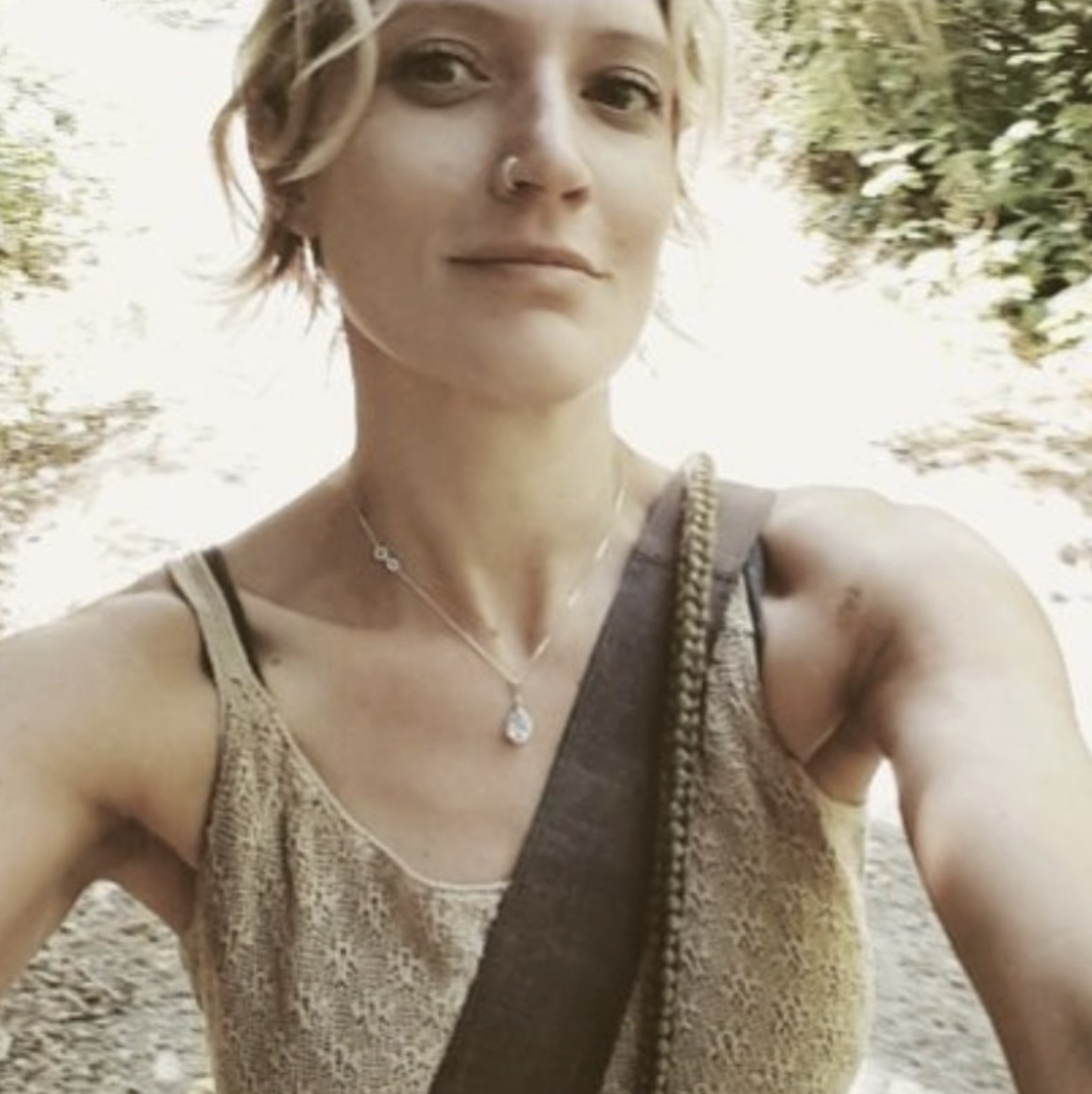 A woman with short blonde hair, wearing a tank top, necklace, and carrying a shoulder strap, taking a selfie outdoors with trees and sunlight in the background.