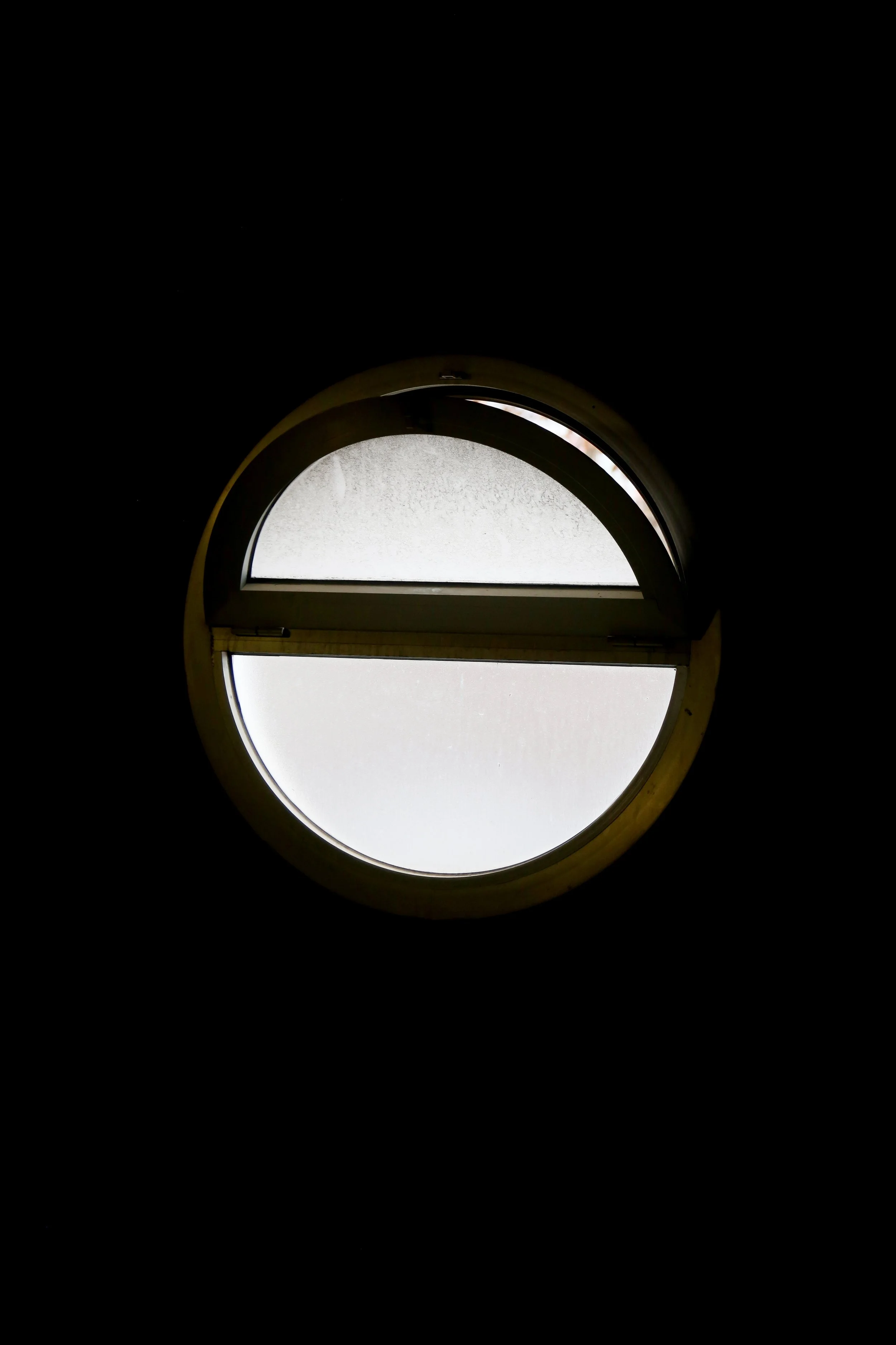 Join us - A circular window with divided upper and lower panes showing frosted or clouded glass, set in a dark wall.