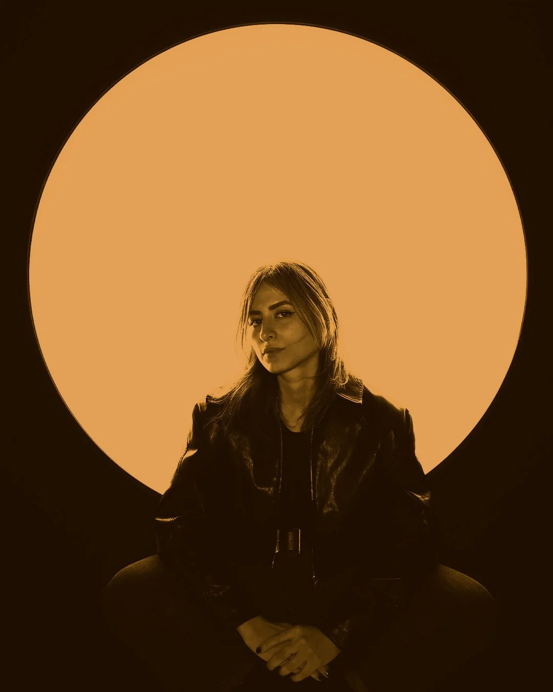 A woman with shoulder-length hair, wearing a black leather jacket, sitting with hands clasped in front of her, against a circular lighted background.
