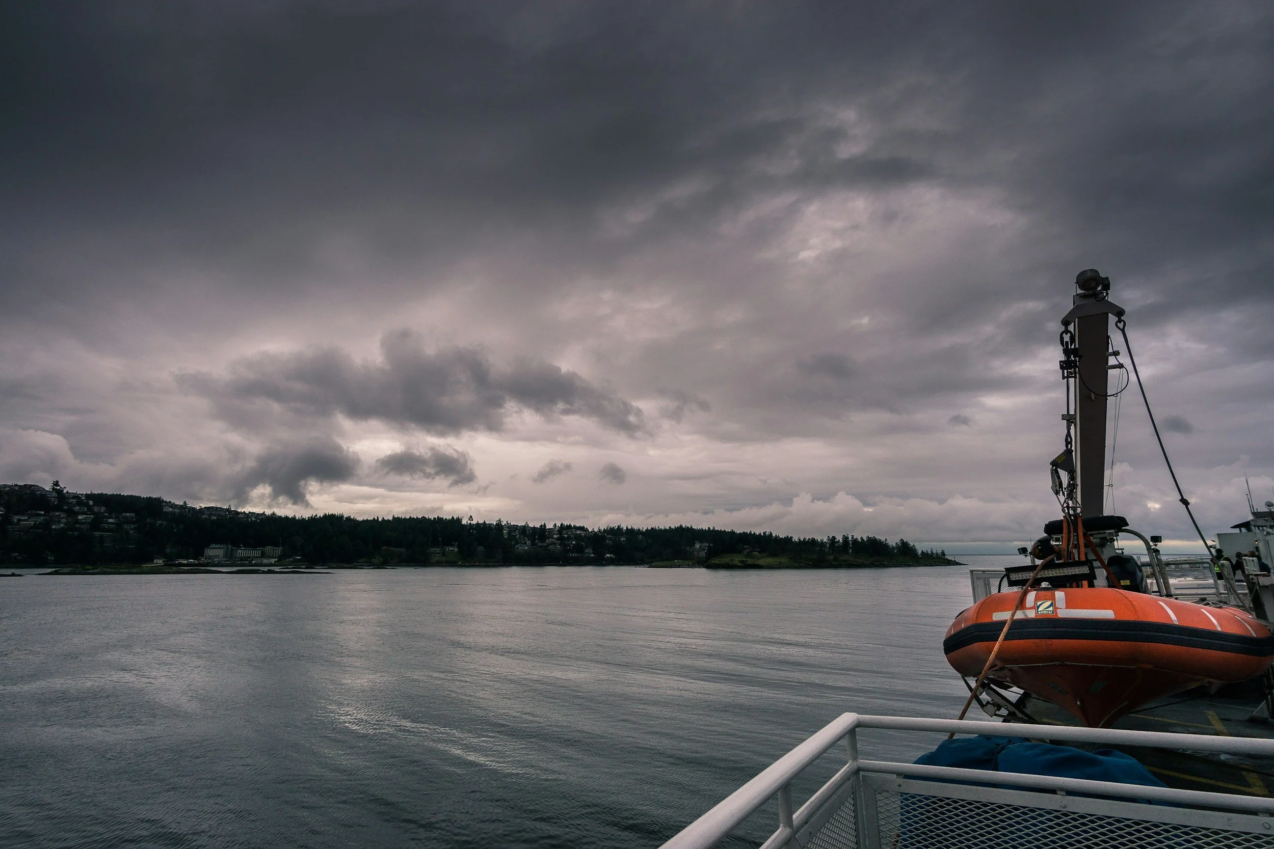 View of a gray, overcast sky over a large body of water with a shoreline and trees in the distance; a bright orange life raft on a boat with a black mast and equipment.