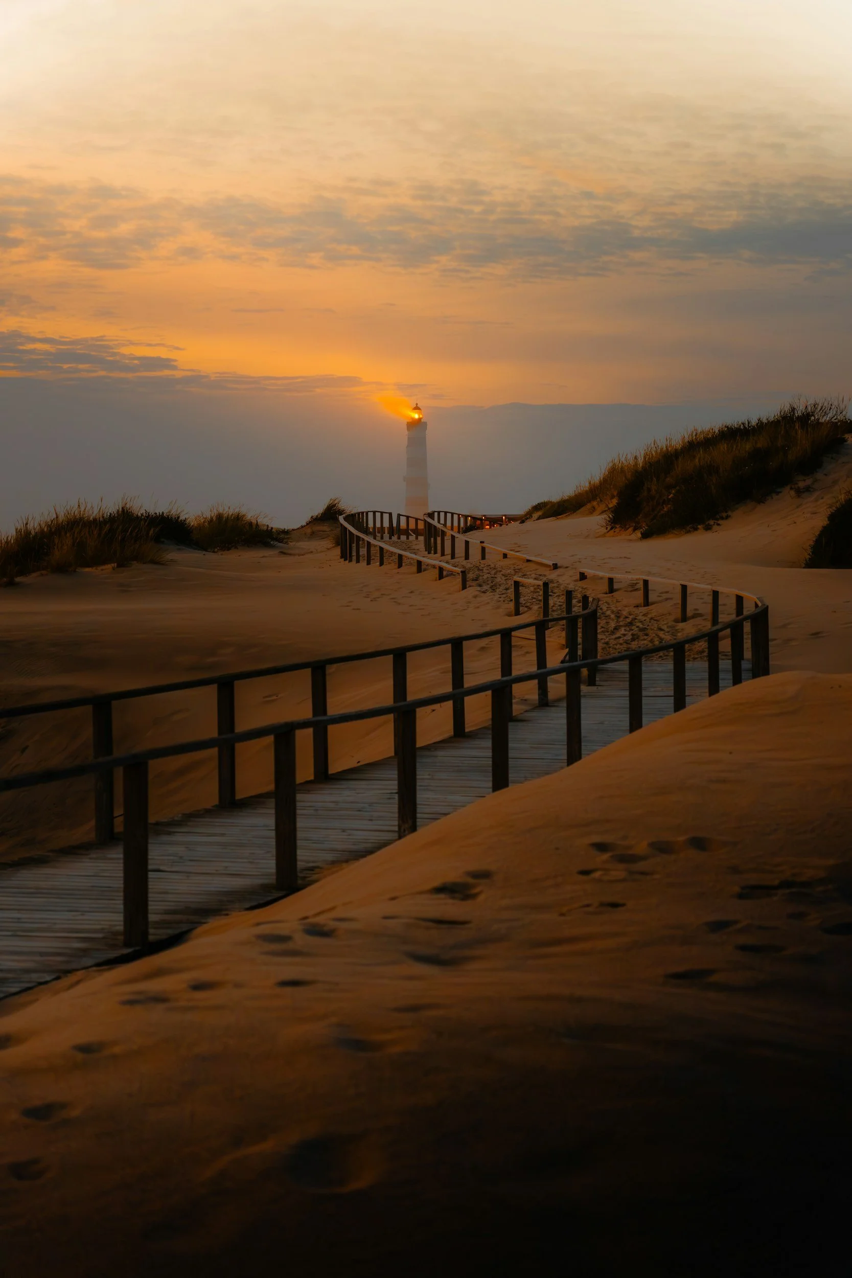 Sand dunes with a wooden walkway leading to a lighthouse during sunset, with low clouds and footprints in the sand.