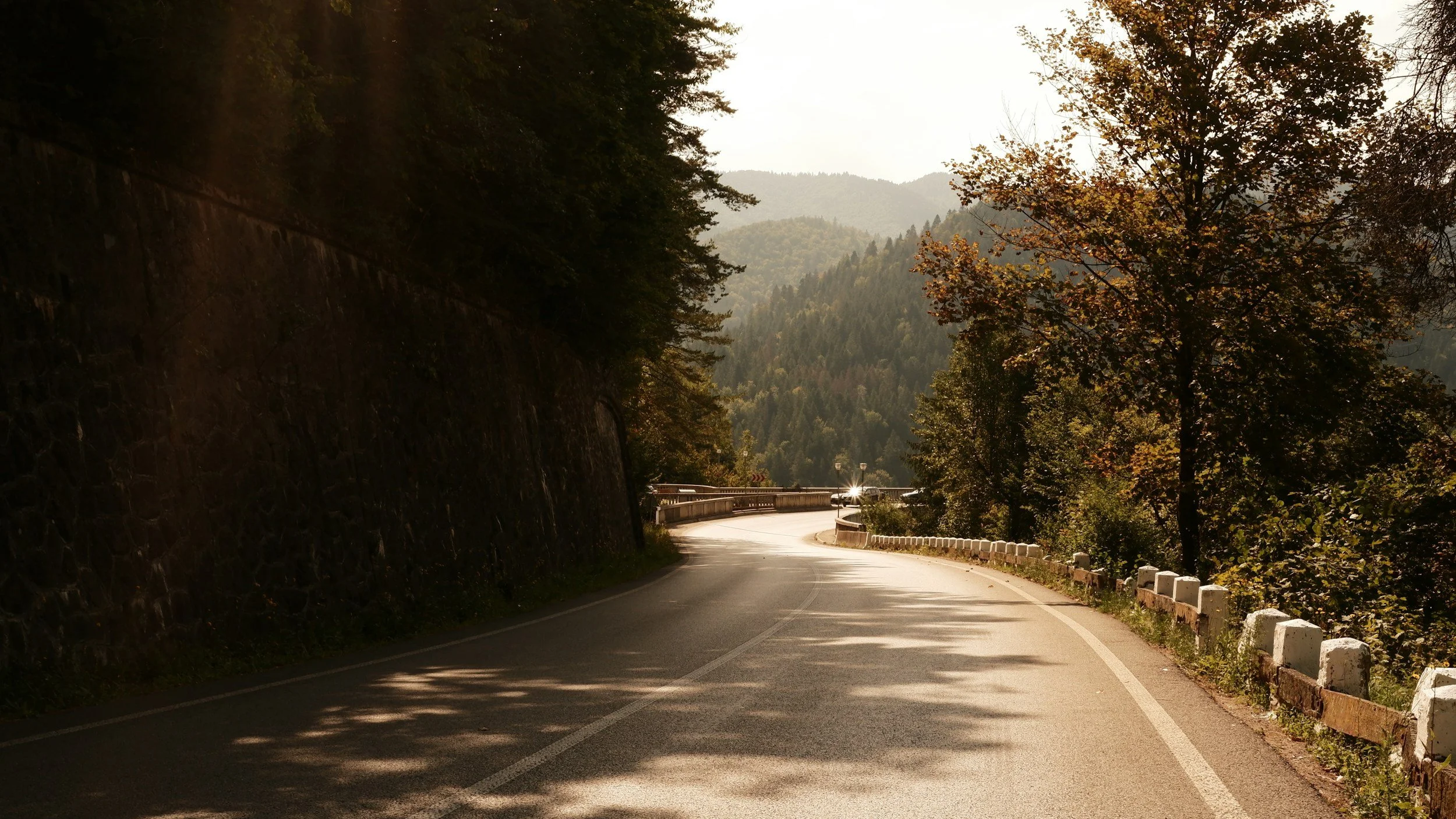 A winding mountain road with guardrails on one side, surrounded by trees with autumn foliage, and distant hills in the background, illuminated by sunlight.