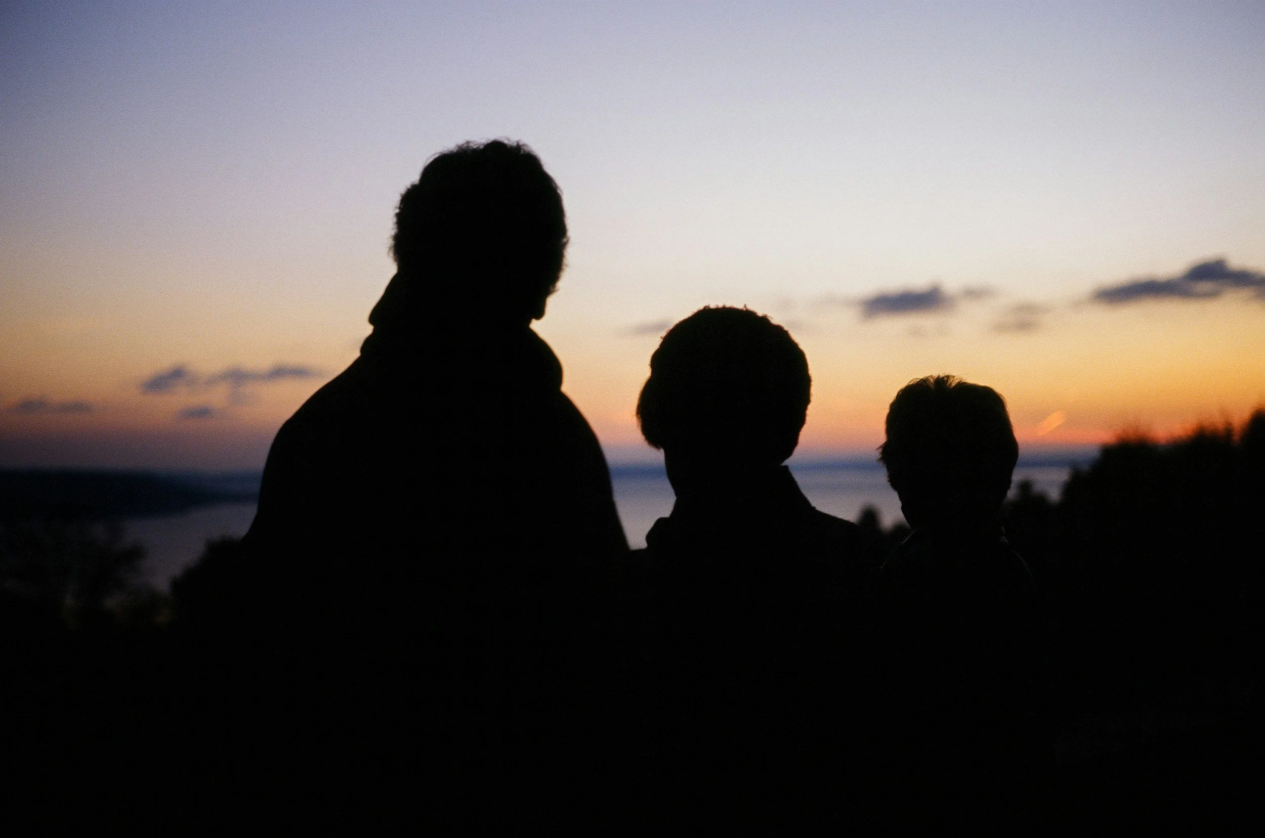 Silhouettes of three people standing outdoors during sunset with a sky and horizon in the background.