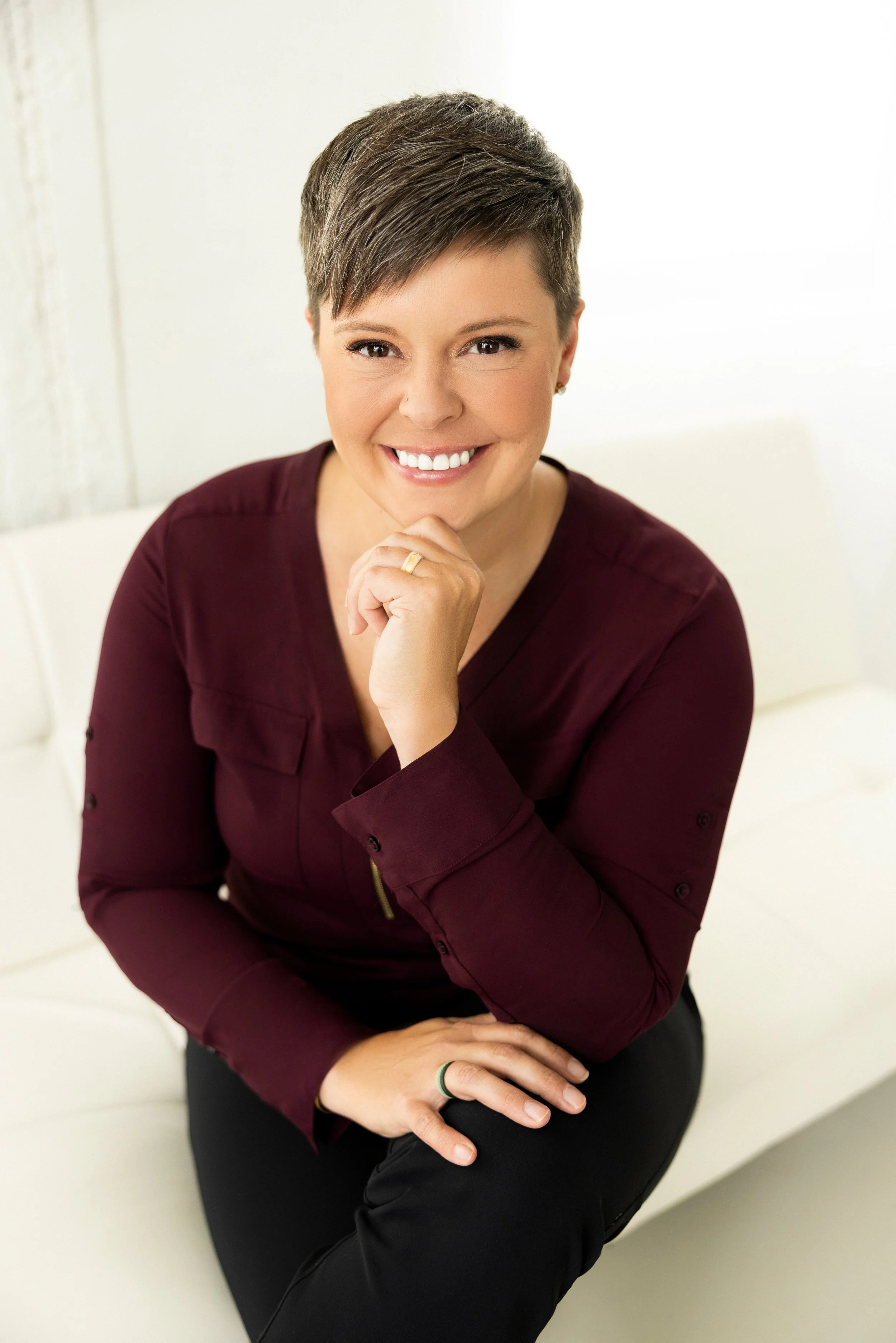 A woman with short hair, wearing a burgundy blouse, sitting on a white couch, smiling, with her chin resting on her hand.