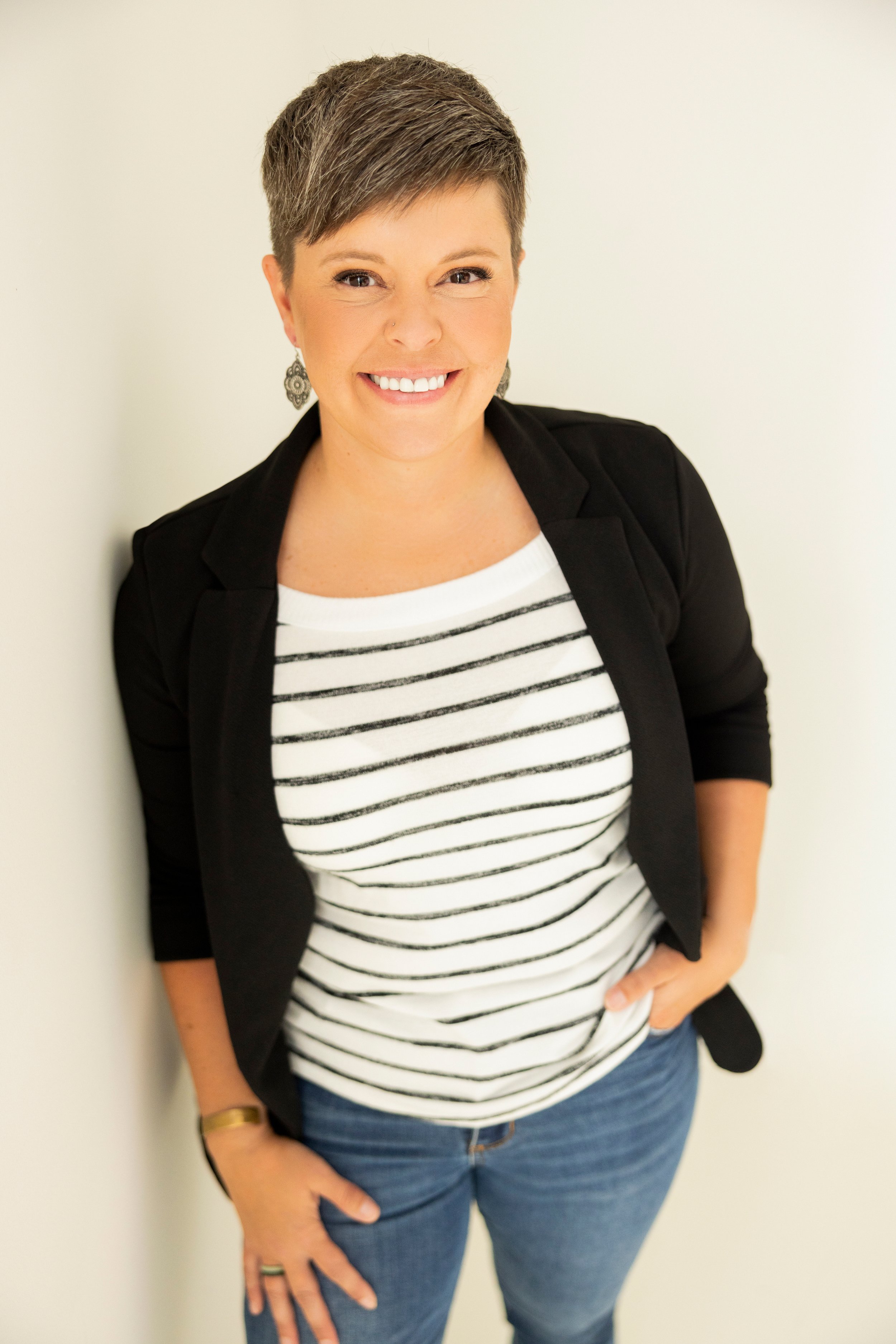 A woman with short hair, wearing a black blazer over a striped shirt, and jeans, smiling while standing against a white wall.