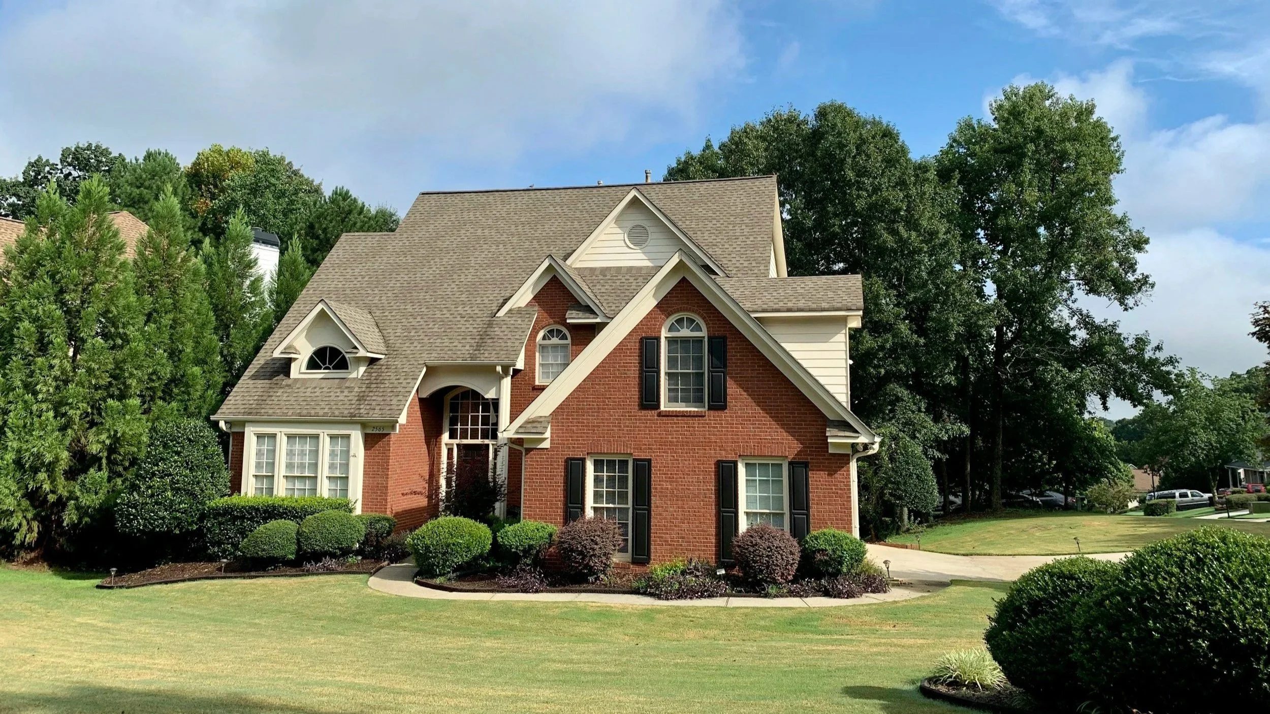 A two-story brick house with a gable roof, surrounded by a well-maintained lawn and landscaped bushes, with tall trees and a partly cloudy blue sky in the background.