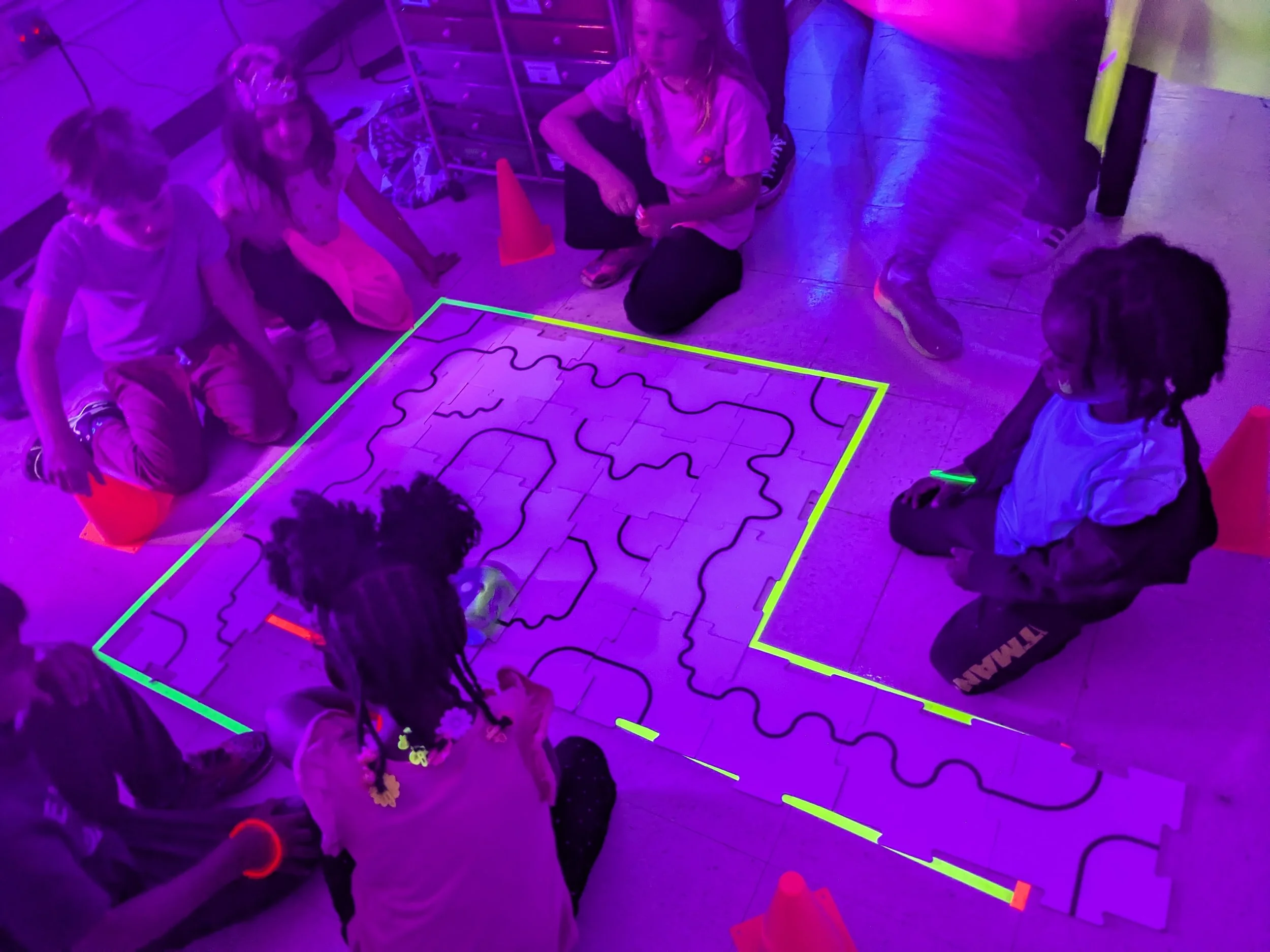 Children gathered around a illuminated maze on the floor with neon lighting, some sitting and some kneeling, and traffic cones placed at corners.