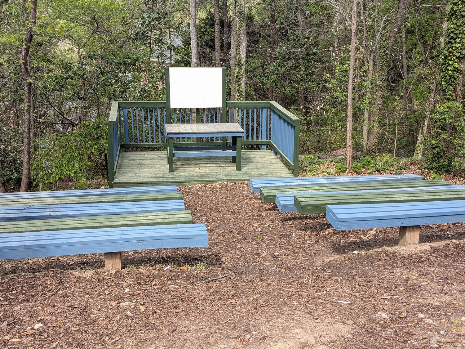 An outdoor scene with tiered blue and green park benches on dirt ground, and a wooden platform with a small table and a whiteboard or sign at the back, surrounded by trees and bushes.
