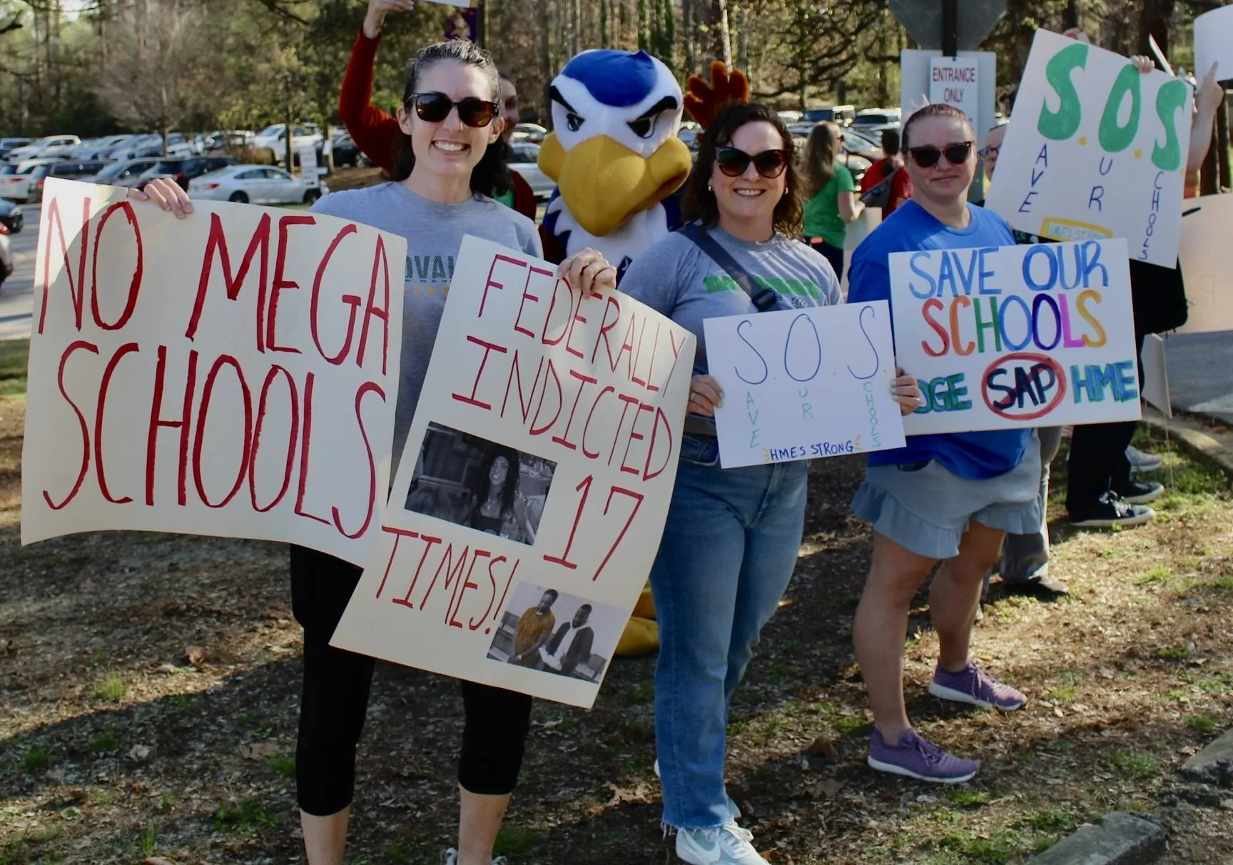 Group of people protesting for education funding, holding signs with messages like 'No Mega Schools,' 'Federally Indicted 17 Times,' and 'Save Our Schools.' One person is dressed in a mascot costume of an eagle, standing among the protesters outdoors near a parking lot.