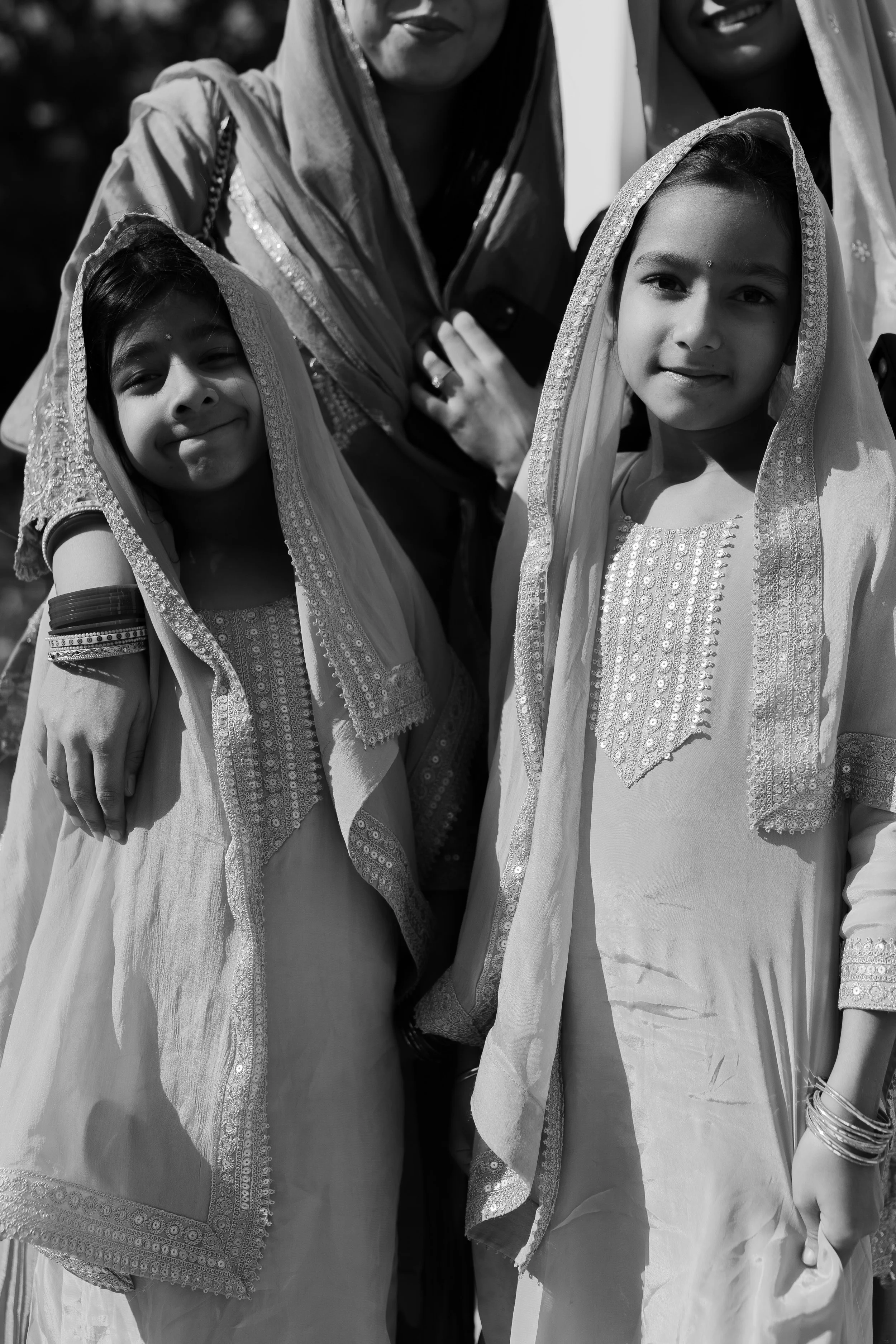 Two young girls and two women wearing traditional Indian clothing and headscarves, standing outdoors in sunlight.