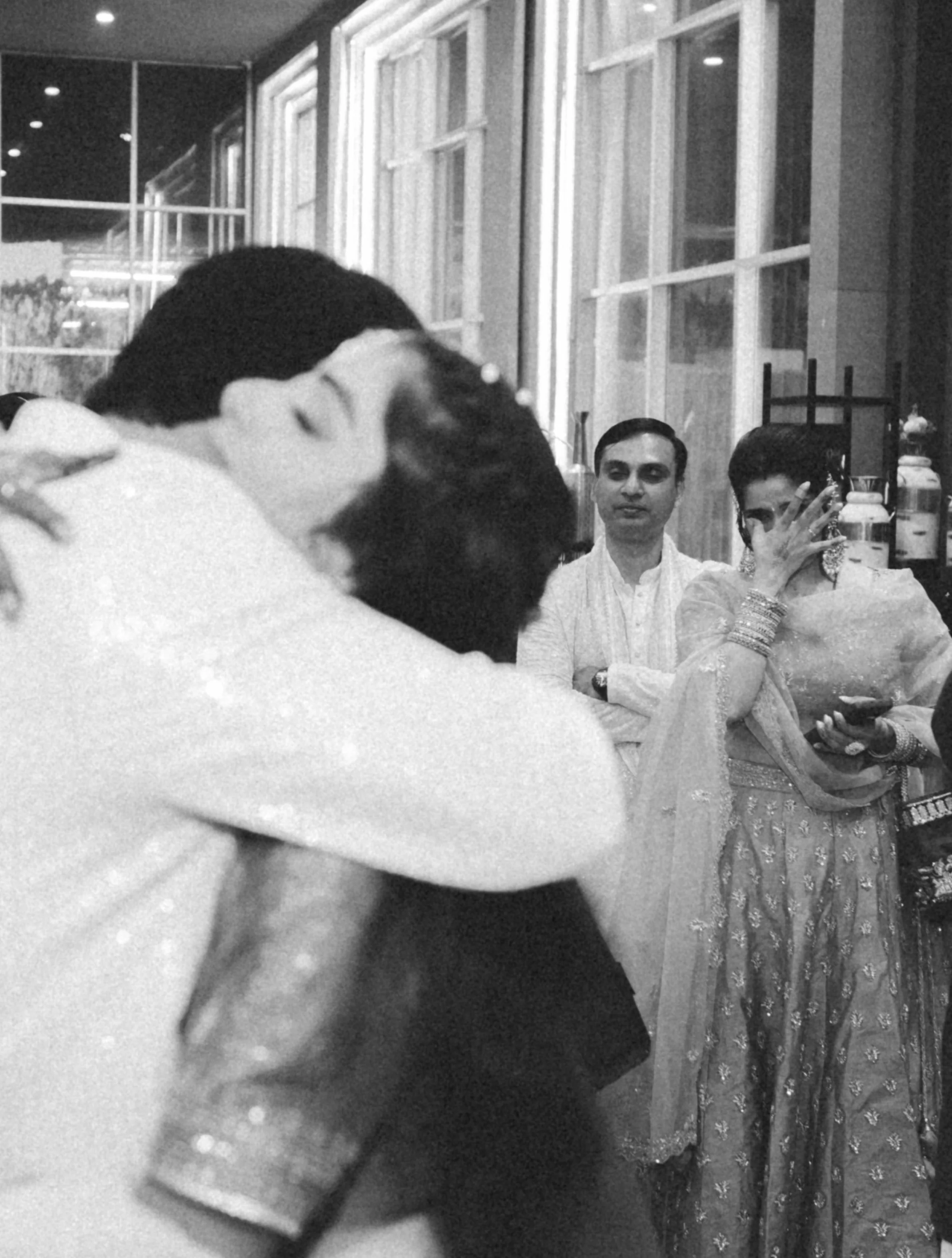 A couple is dancing closely at an event, with bystanders watching. One woman is covering her face with her hand, and two men are standing behind them, observing. The woman is dressed in a traditional outfit with jewelry, and the man is wearing a ligh