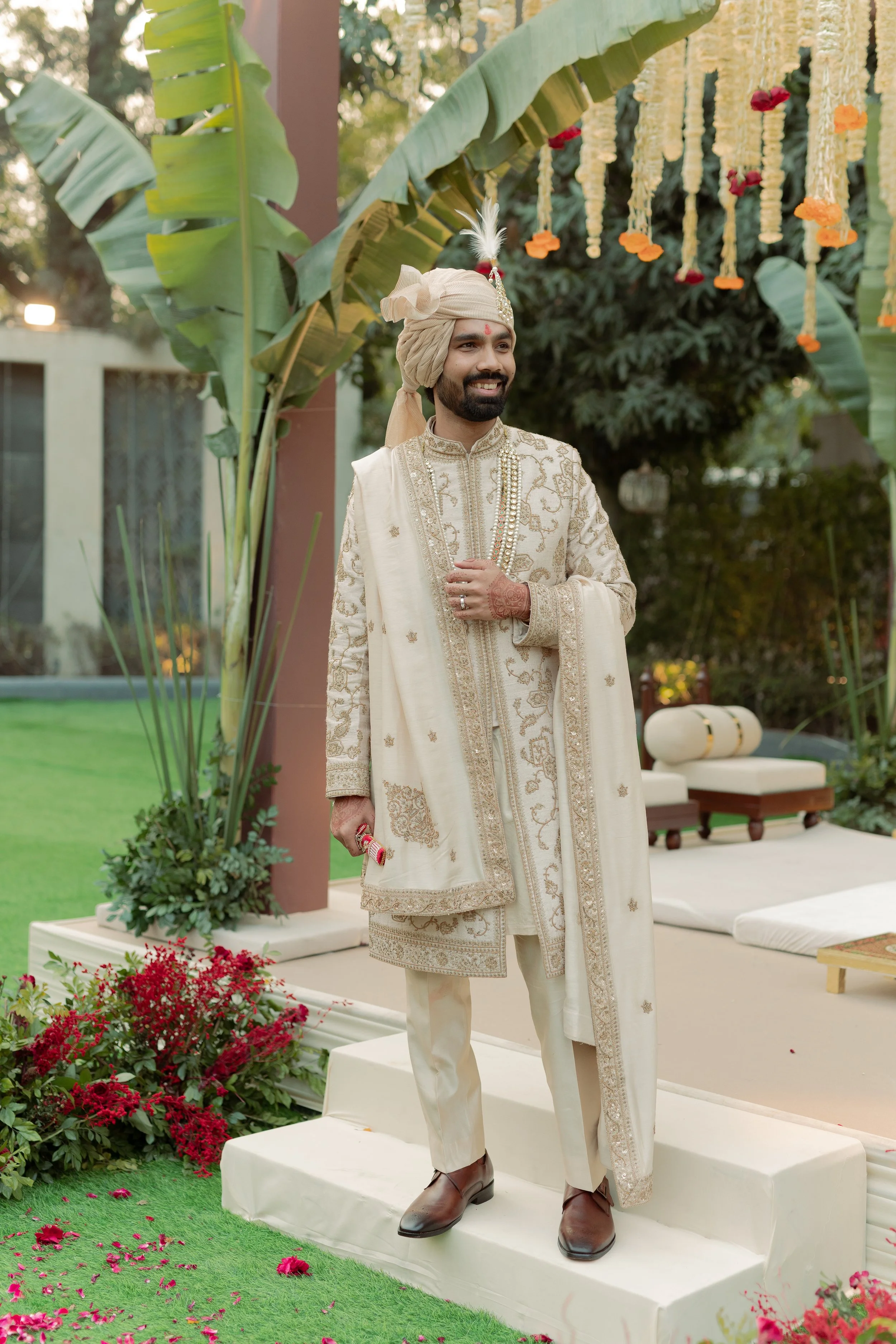 A man dressed in traditional Indian wedding attire standing on a white platform in an outdoor decorated garden with green leaves, flowers, and hanging marigold garlands.