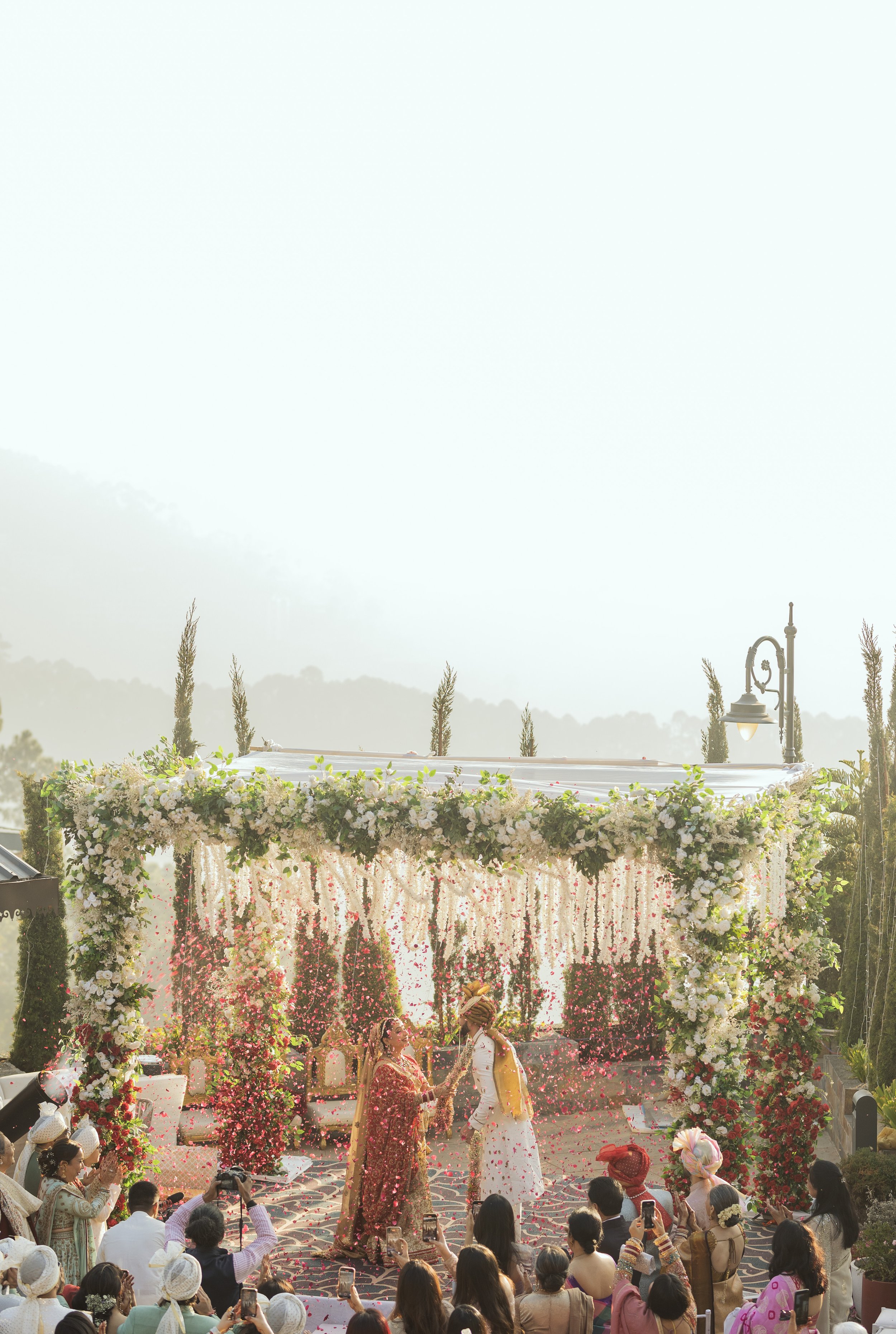 Outdoor wedding ceremony with floral arch and guests, two people exchanging rings, confetti falling, sunny day.