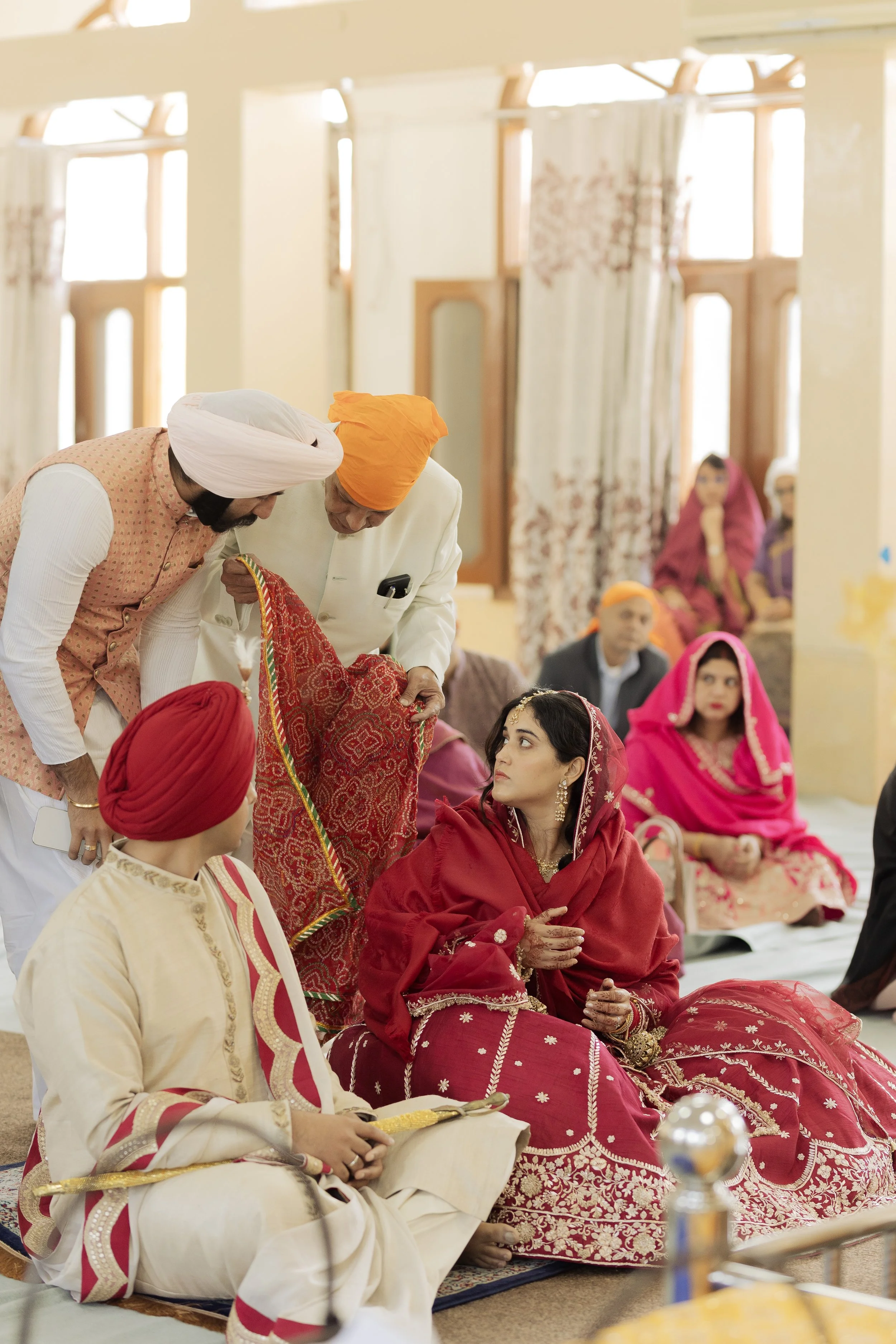 Indian wedding ceremony with women dressed in traditional red and pink bridal attire and men in colorful traditional clothing, inside a decorated venue.