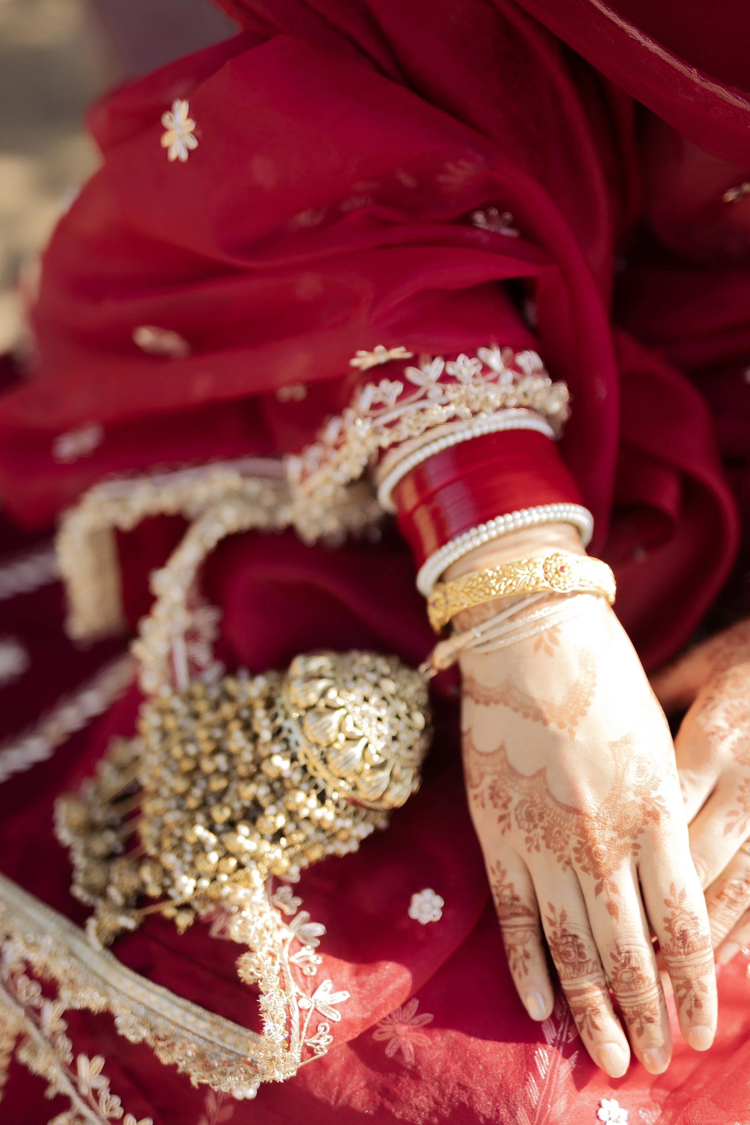 Close-up of a woman's hand adorned with mehndi and jewelry, resting on red and gold fabric with traditional Indian jewelry, including bangles and a large necklace.