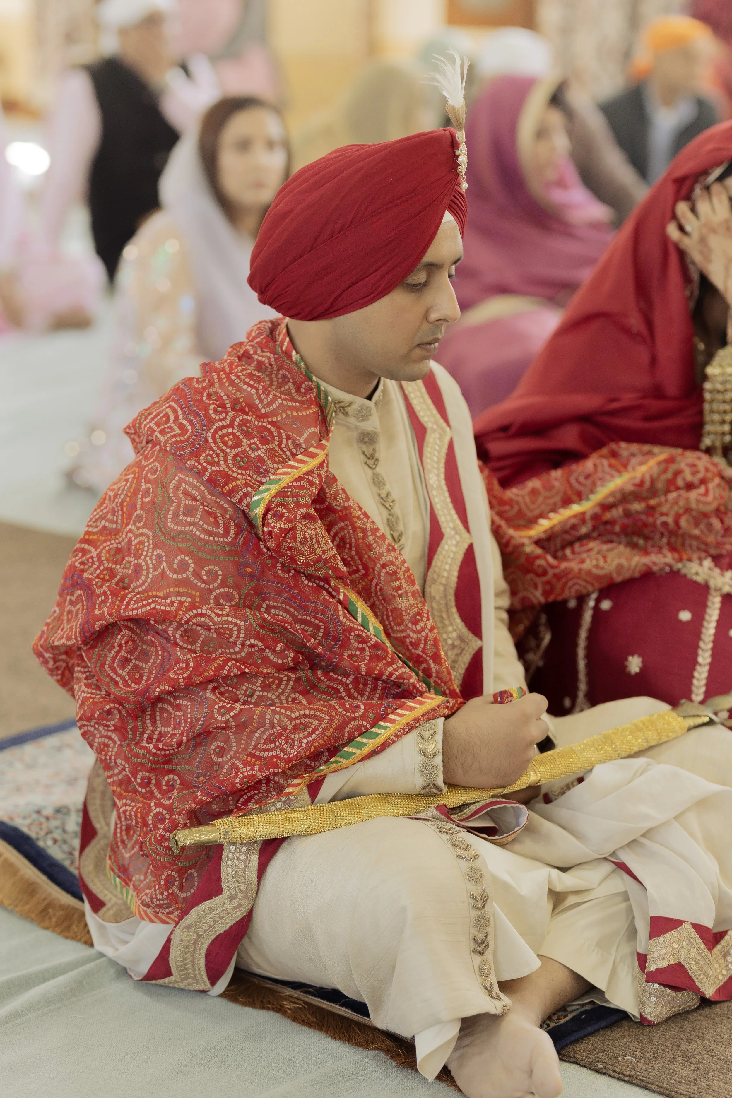 A man dressed in traditional South Asian wedding attire, including a red turban with a feather, cream-colored kurta, and embroidered shawl, participating in a wedding ceremony.
