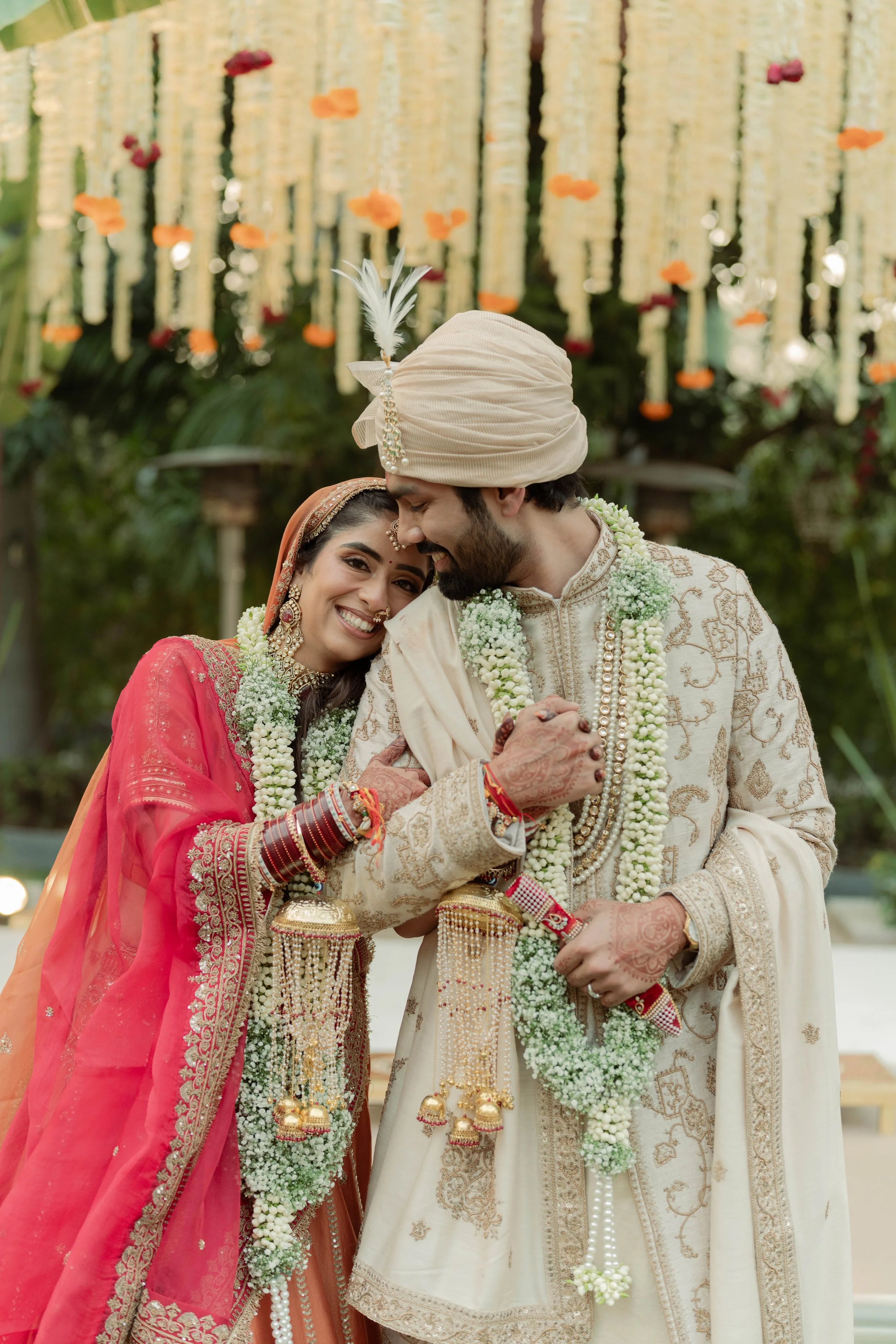 Indian wedding couple dressed in traditional attire, smiling and embracing, with decorative backdrop of marigold flowers and hanging floral garlands.