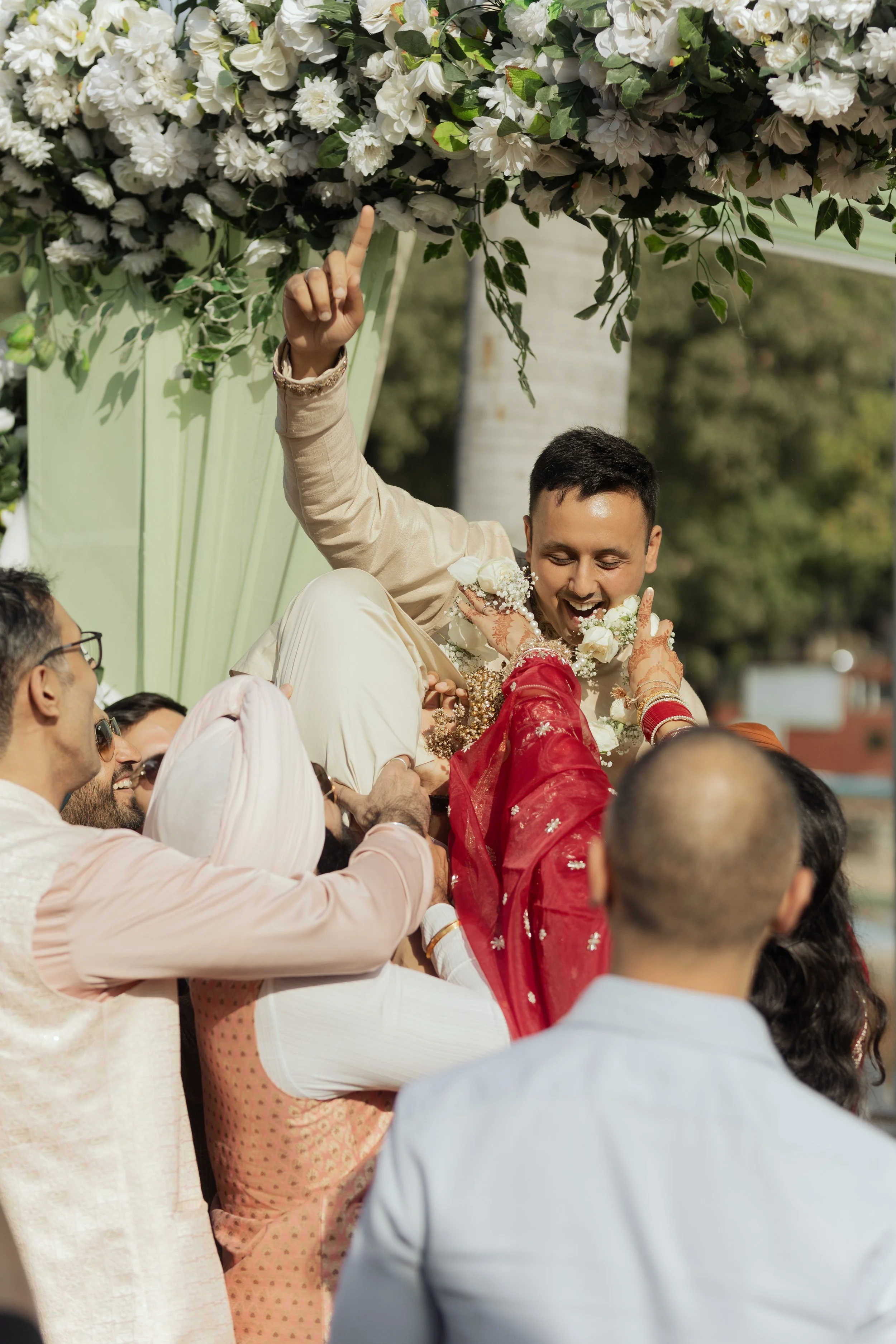 A groom being lifted by friends during a traditional Indian wedding celebration, with floral decorations above them.