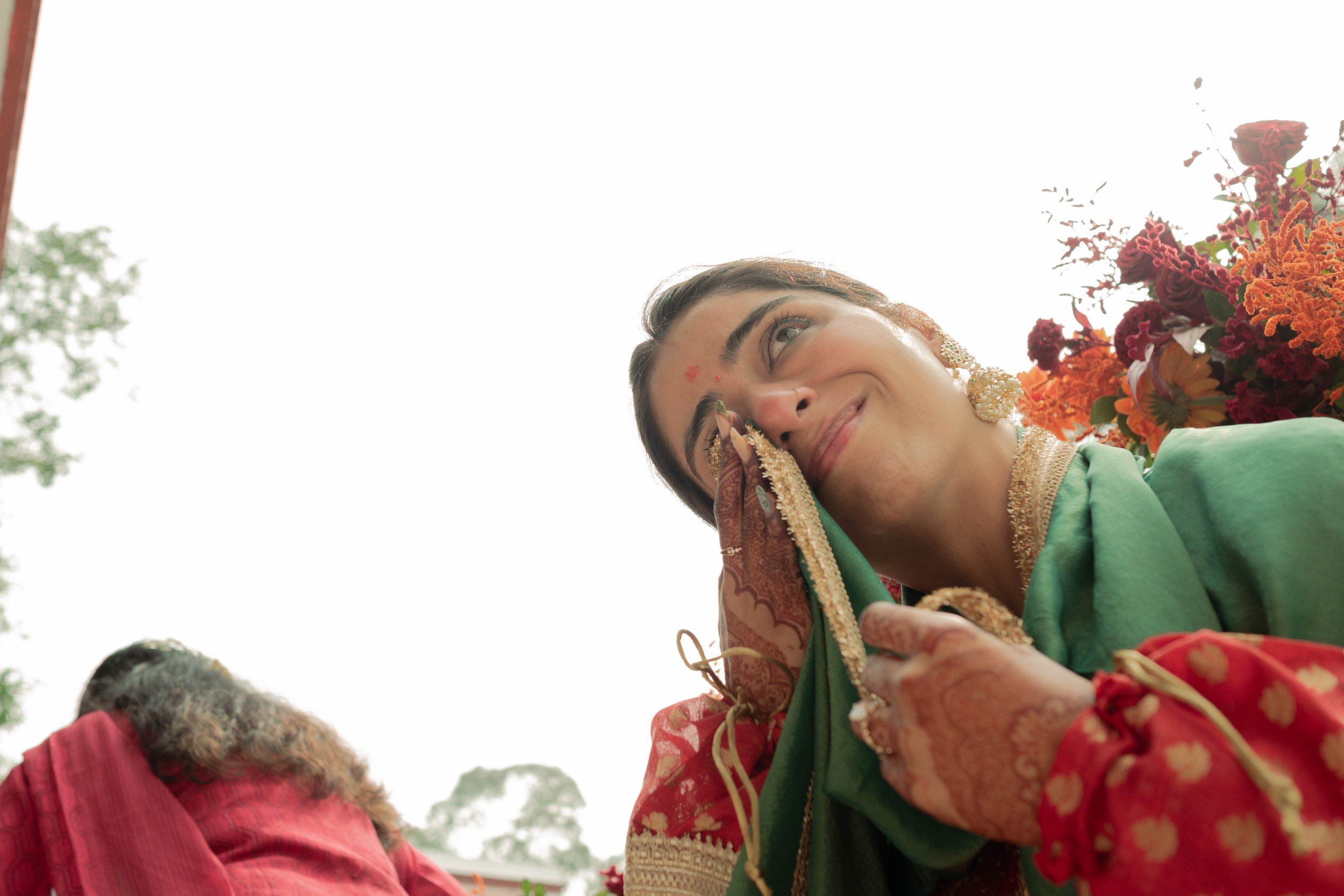 A woman in traditional Indian attire smiling and touching her face, surrounded by colorful flowers, participating in an outdoor cultural or religious celebration.