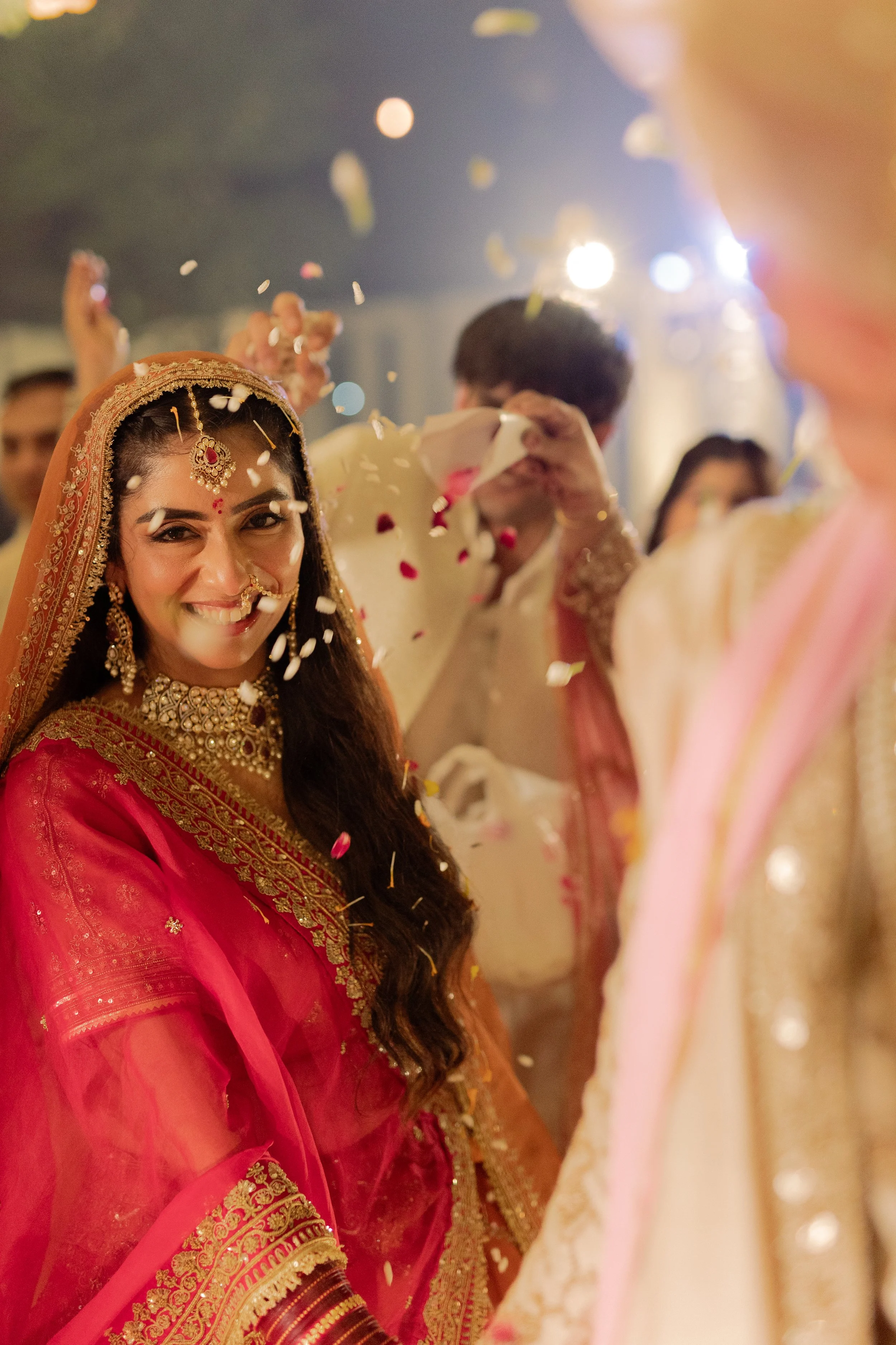 A woman in traditional Indian wedding attire smiling as flower petals are showered on her during a celebration.