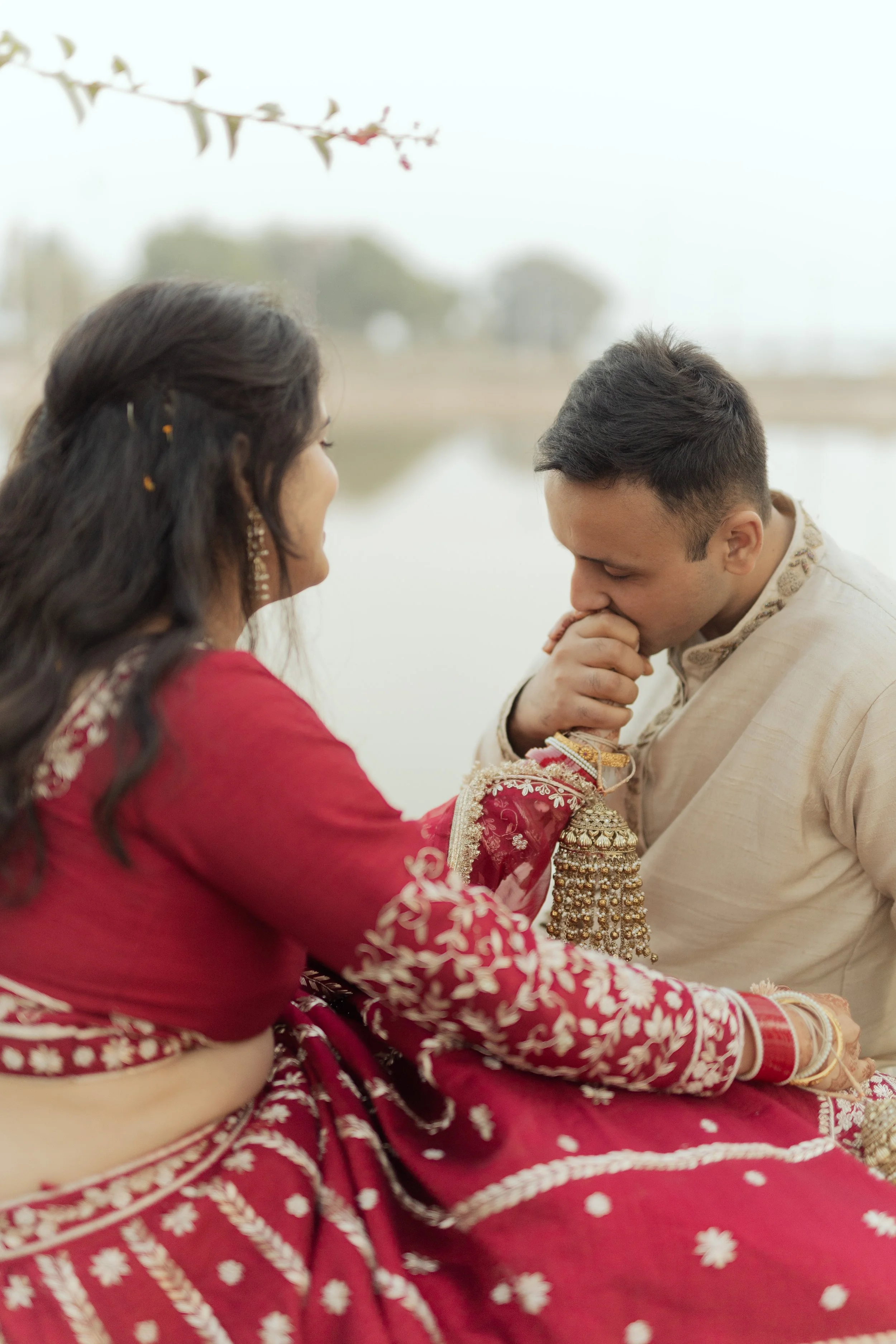 A man kneels and holds a woman's hand near his mouth during a traditional Indian wedding ceremony outdoors by a lake, with trees in the background.