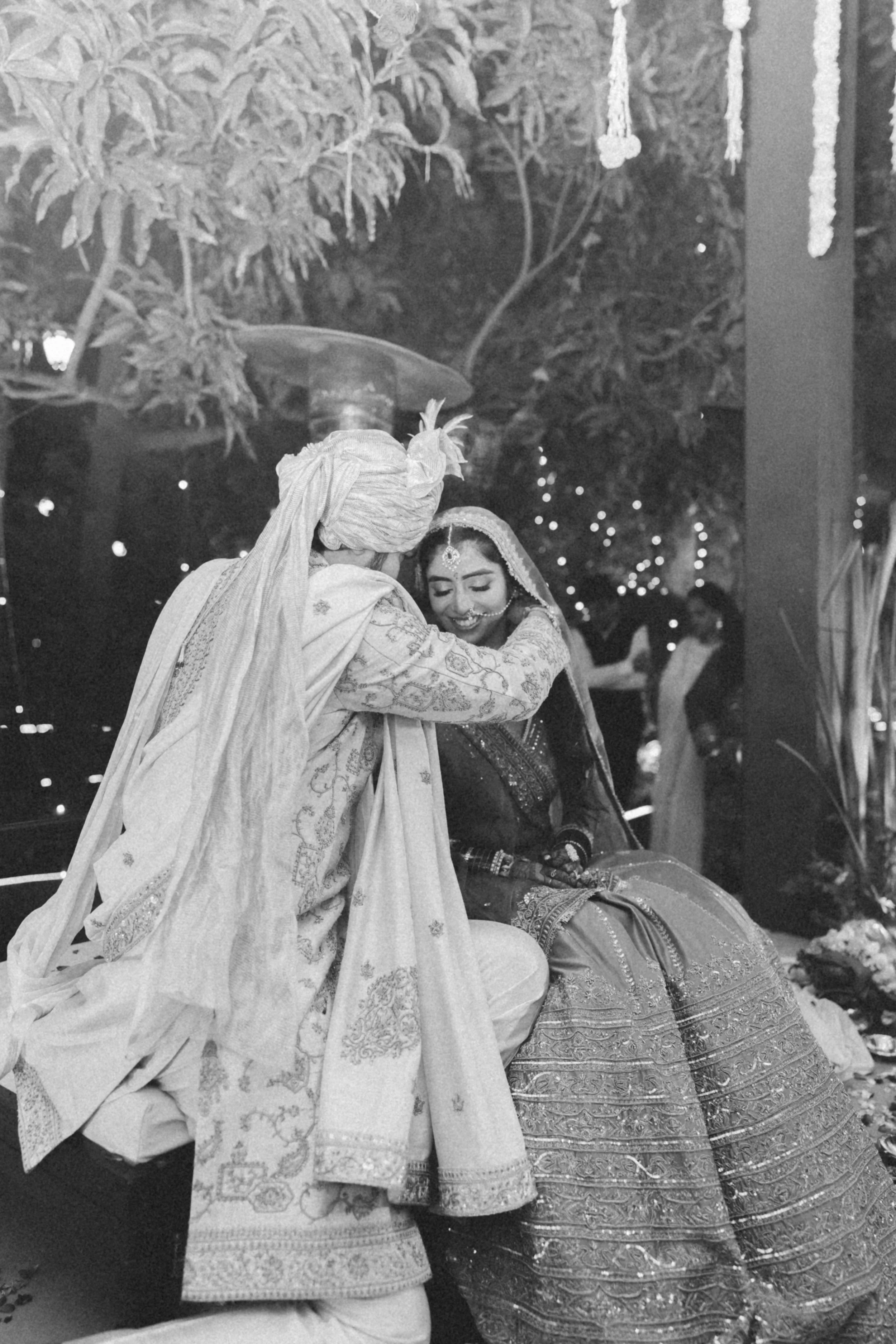 A couple dressed in traditional Indian wedding attire sharing a tender moment, with the groom kissing the bride's forehead, both smiling, in a decorated wedding venue with hanging decorations and string lights.