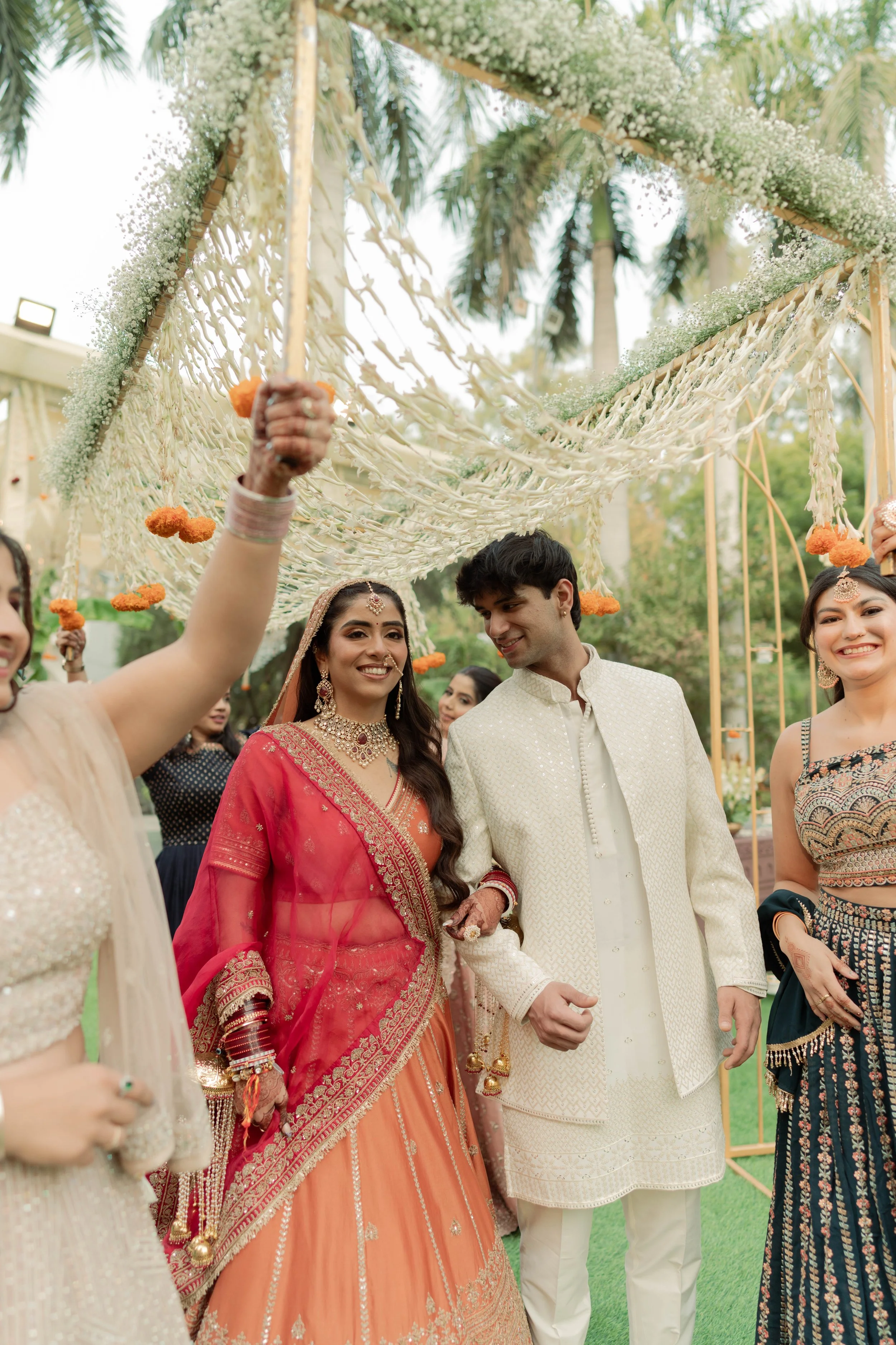 A group of people celebrating at an outdoor Indian wedding, with a bride in a red and gold saree, a groom in a cream-colored sherwani, surrounded by friends holding flower garlands, under a decorated canopy with hanging flowers and greenery.