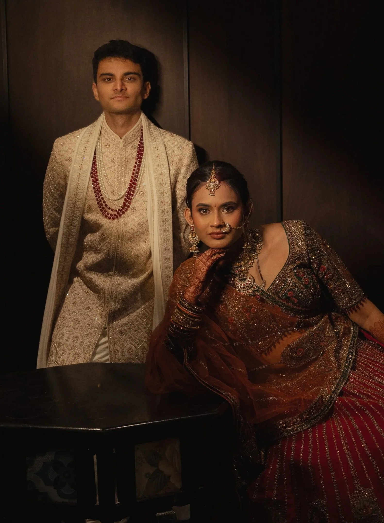 A man and woman dressed in traditional Indian wedding attire, with the man standing and the woman leaning on a table, inside an indoor setting with dark wood-paneled walls.