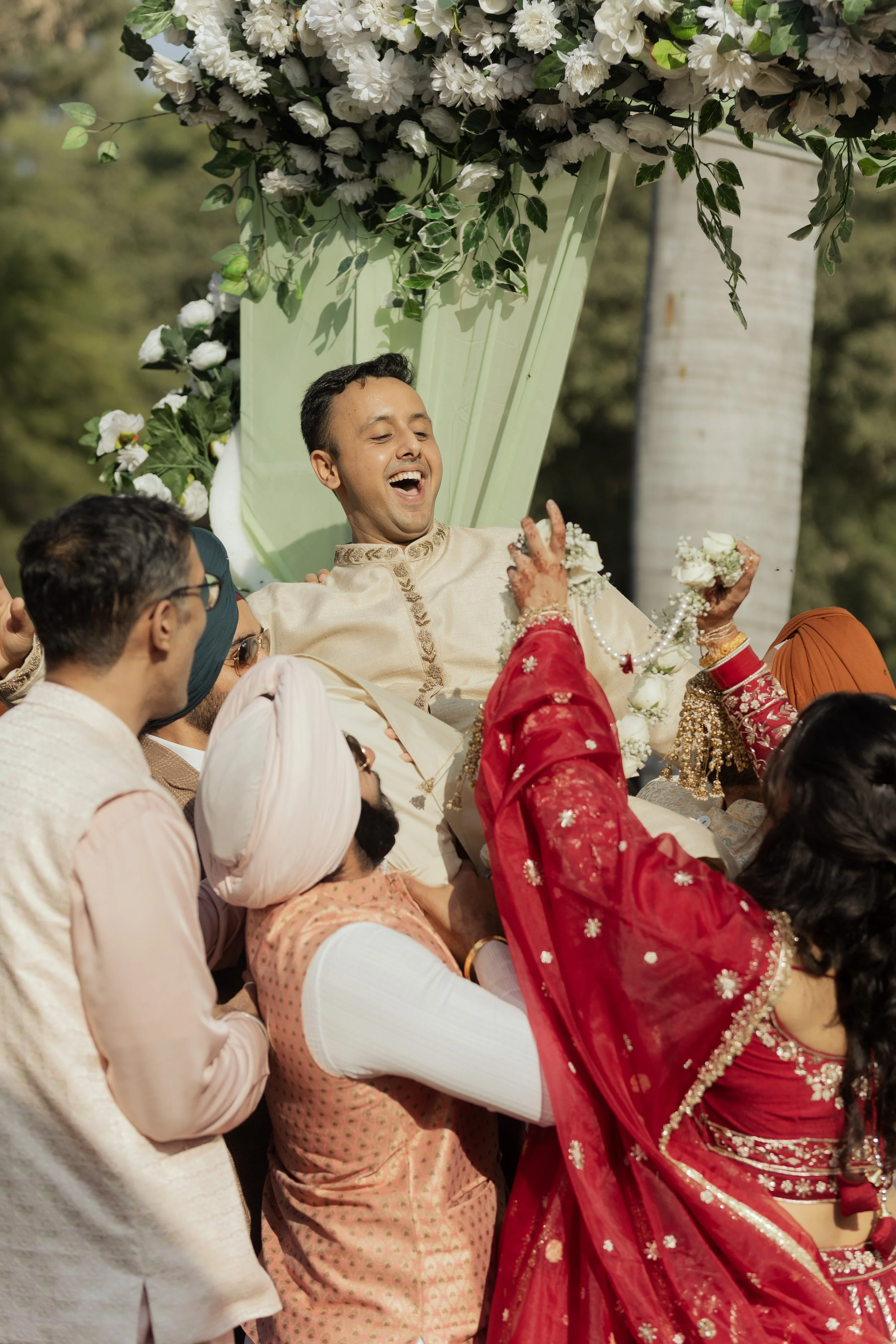 Happy groom being lifted by friends and family during an outdoor wedding celebration, surrounded by floral decorations and trees.