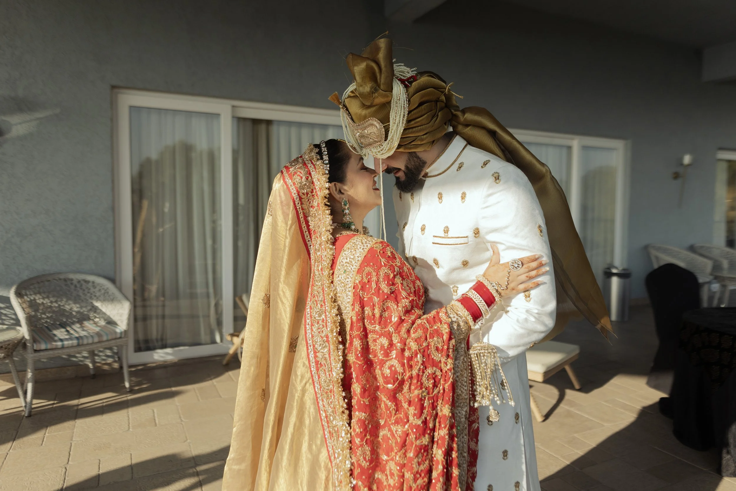 Indian bride and groom in traditional wedding attire sharing a kiss outdoors.