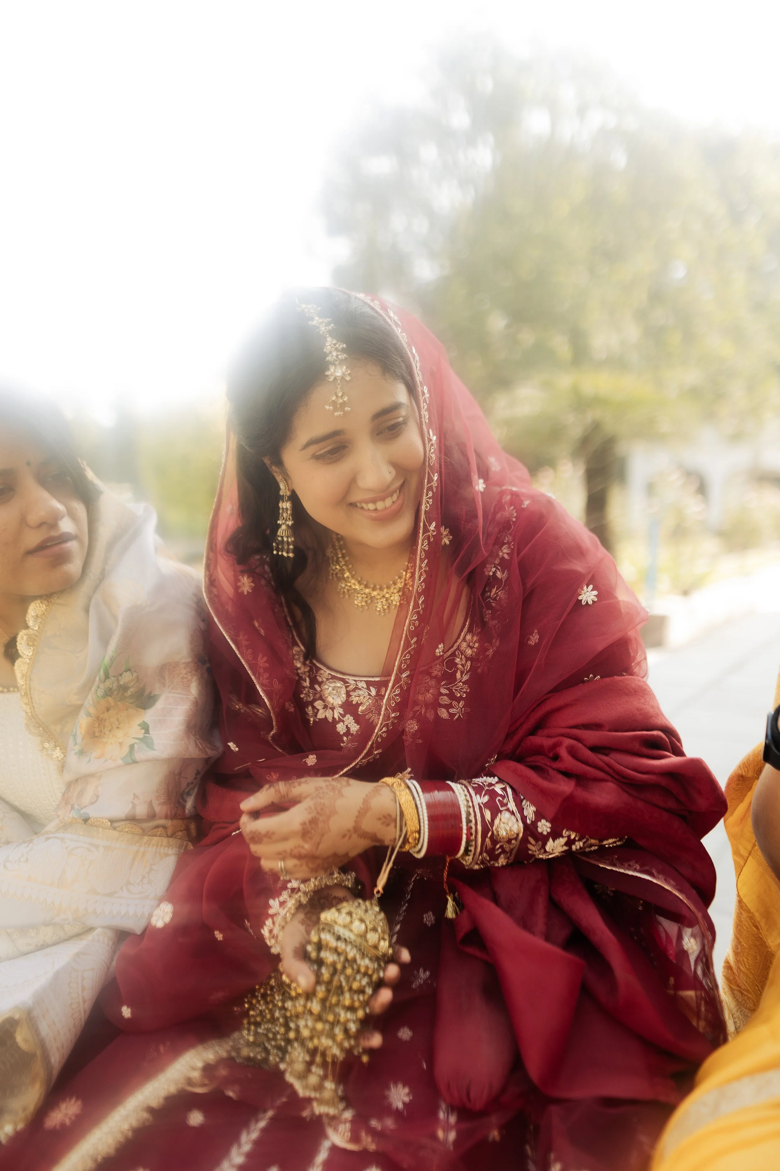 A woman dressed in a red traditional Indian outfit with jewelry, smiling and holding gold-colored accessories, sitting outdoors.
