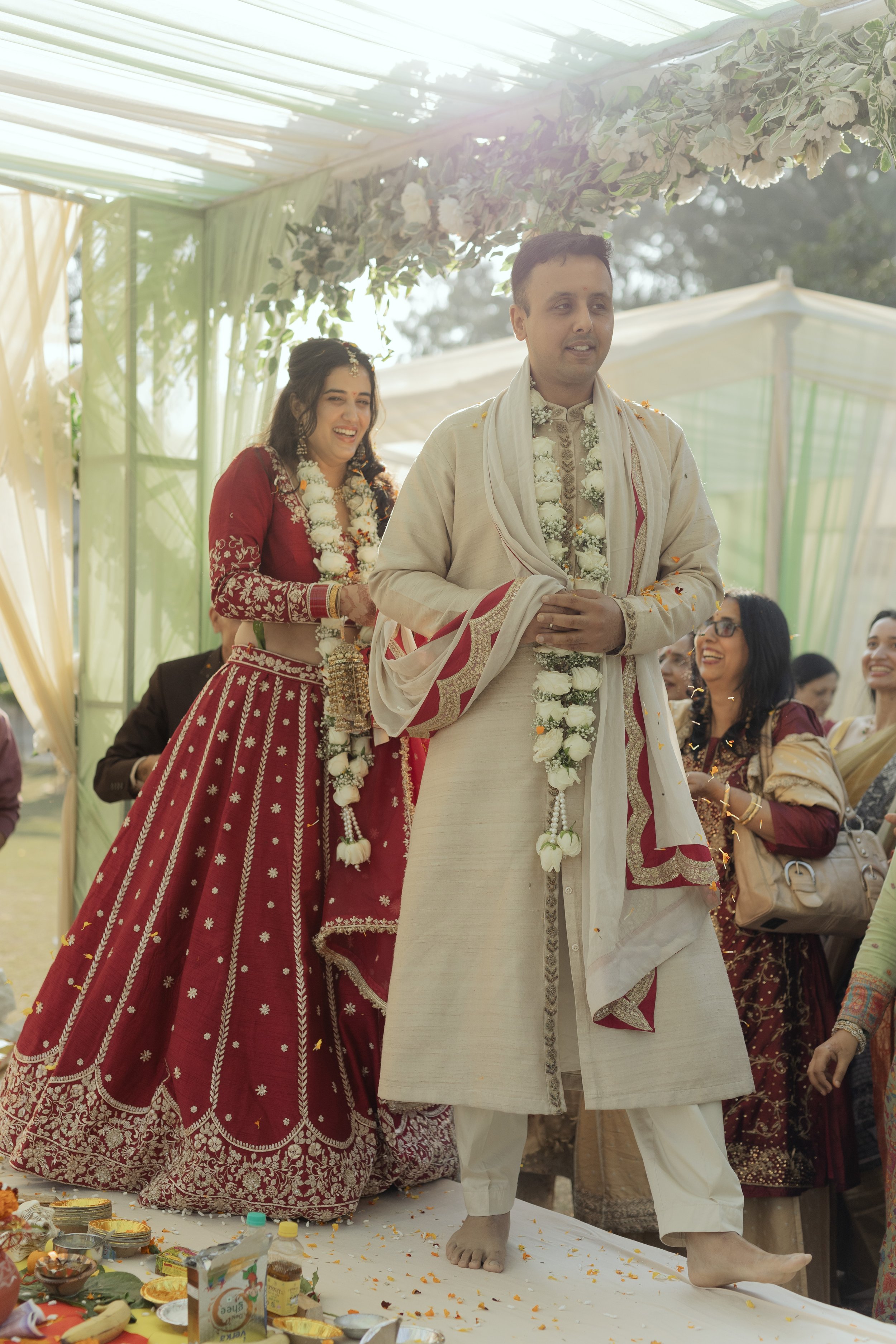 Indian wedding ceremony with bride and groom standing on an altar, wearing traditional attire and floral garlands, surrounded by family and friends.