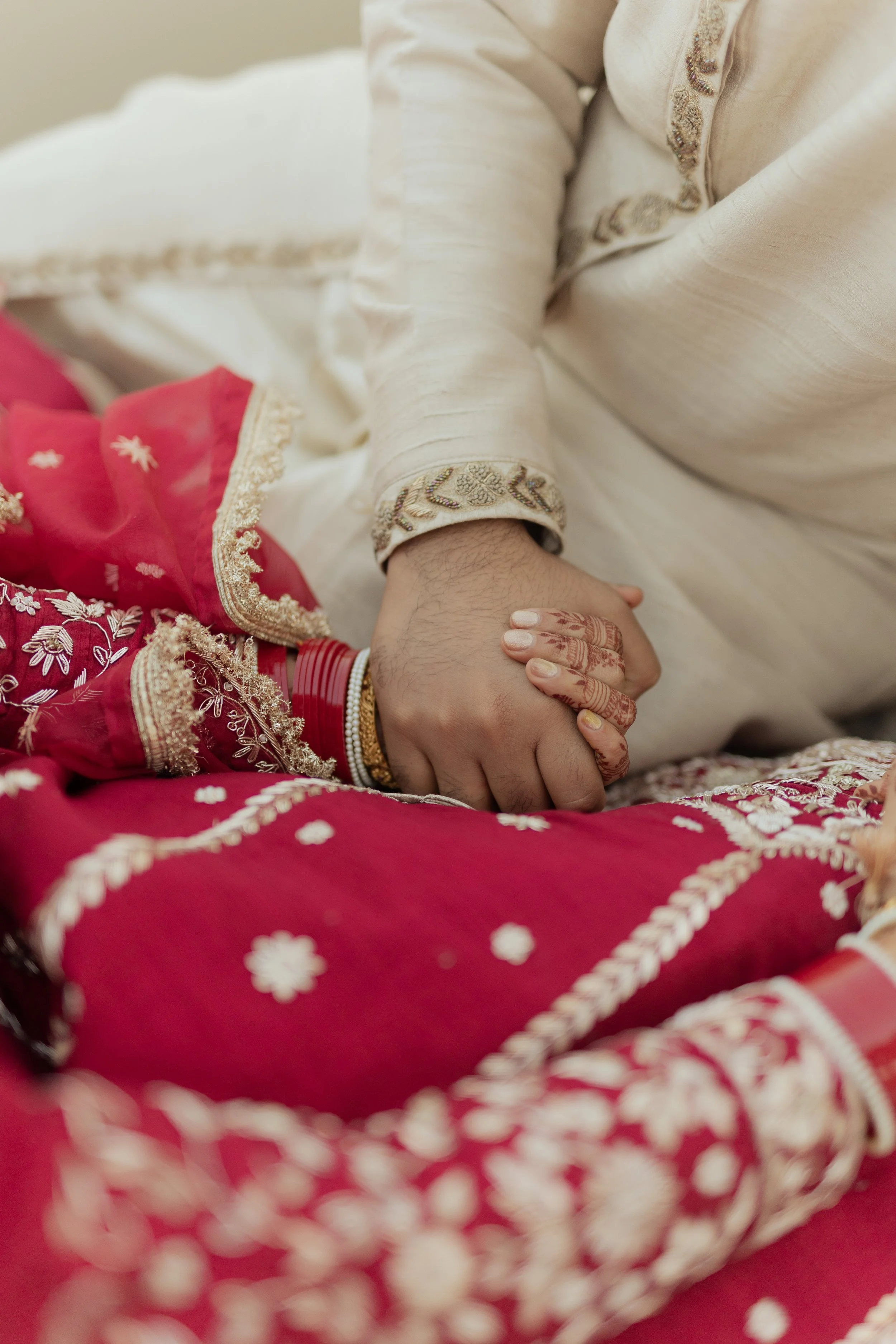 Close-up of a bride and groom holding hands during a traditional Indian wedding ceremony, with intricate henna designs on the bride's hand and colorful embroidered clothing.