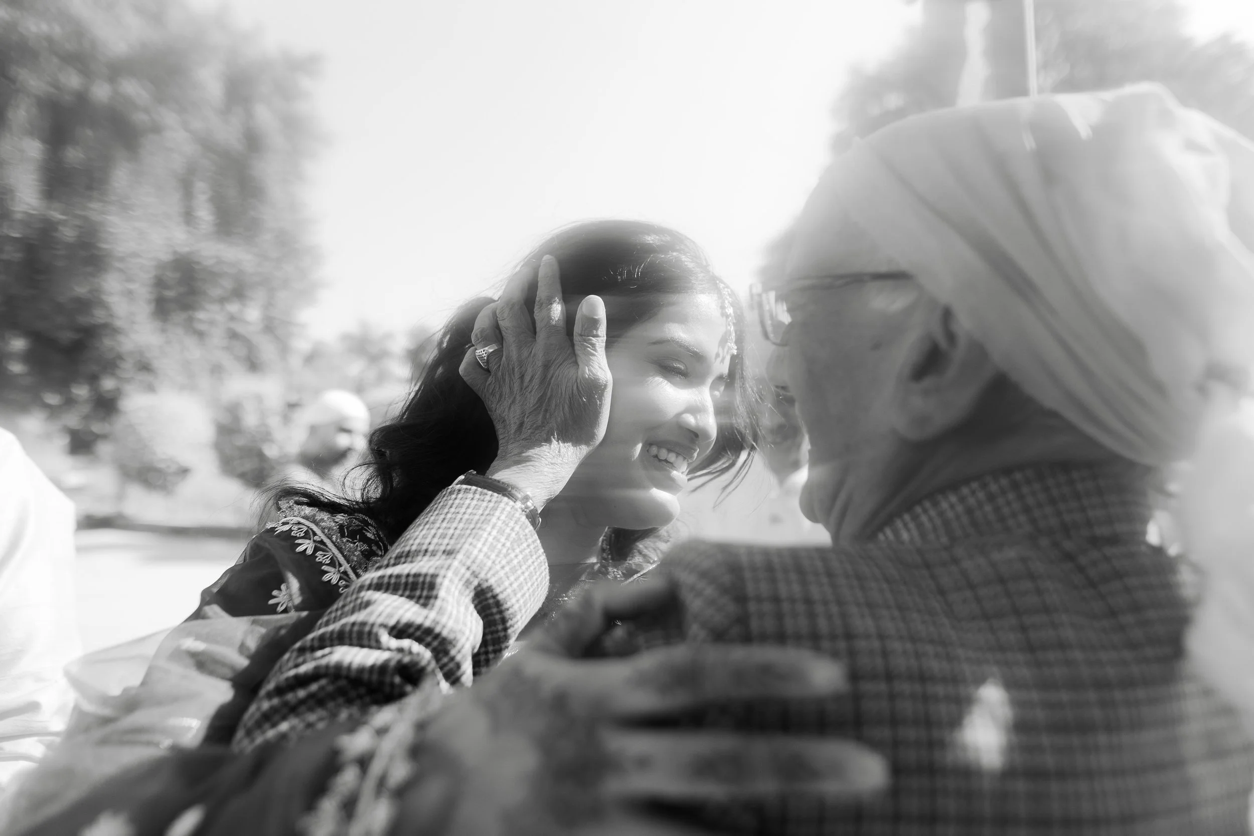 A young woman smiling and gently holding the face of an elderly man in a turban, both outdoors on a sunny day.