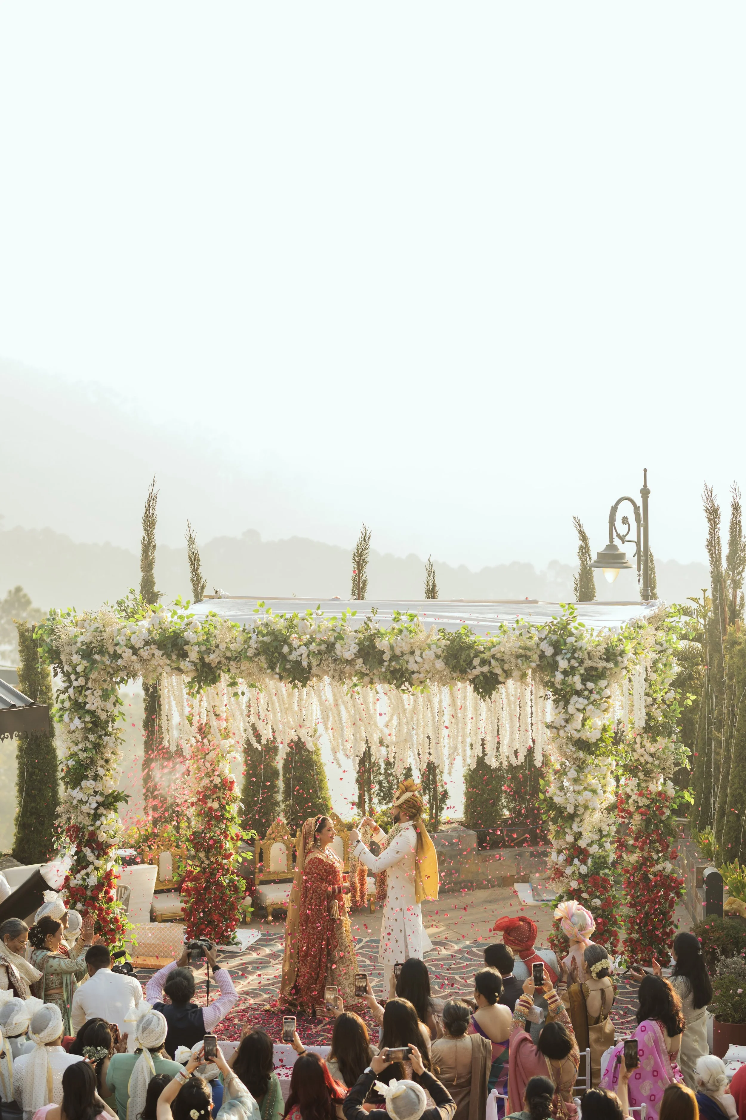 A traditional wedding ceremony with a floral decorated canopy and audience taking photos.