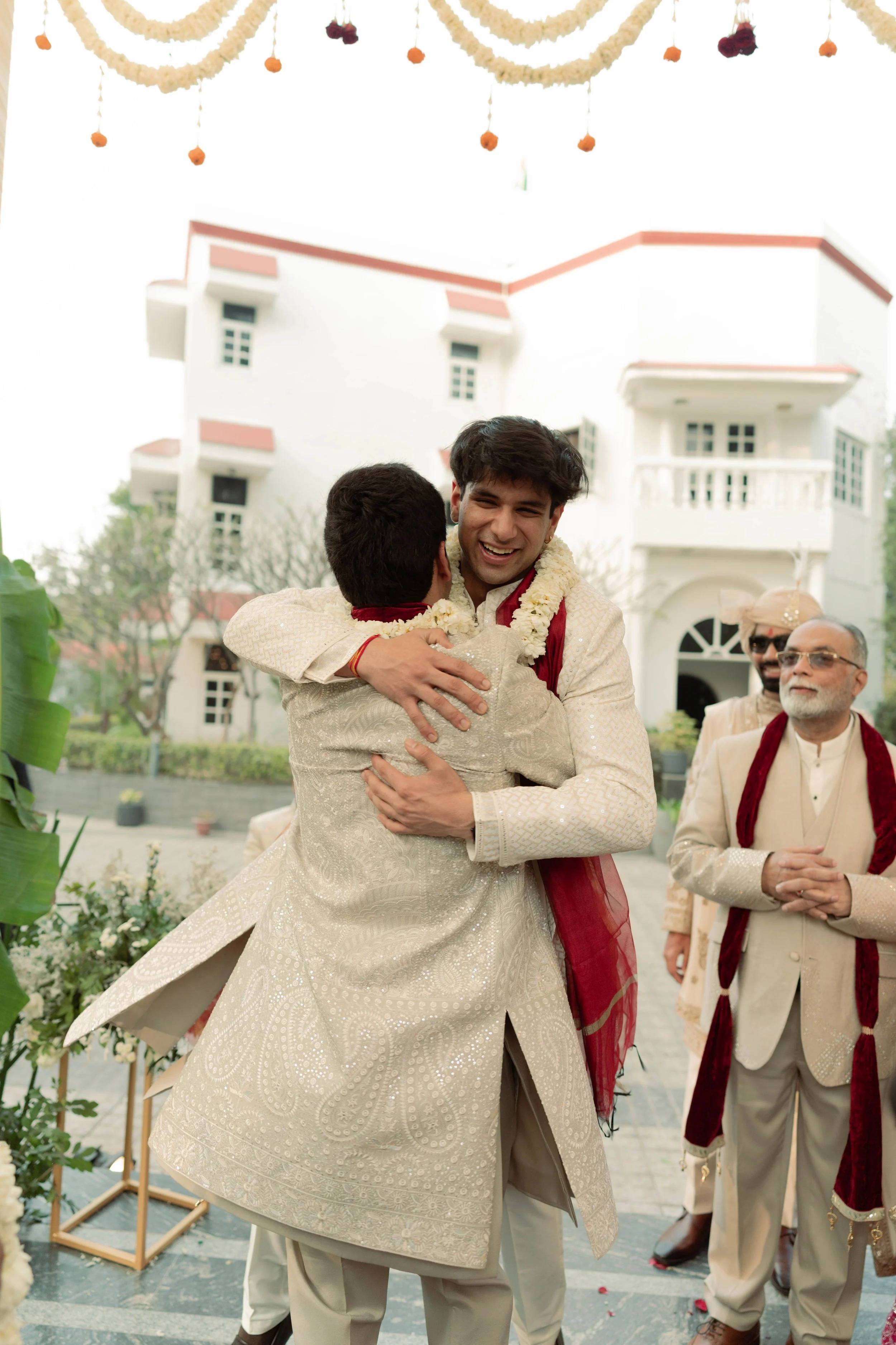 Two men embracing in traditional Indian wedding attire during a celebration, with other men in similar attire standing nearby outside a white building with balconies, decorated with hanging flower garlands.