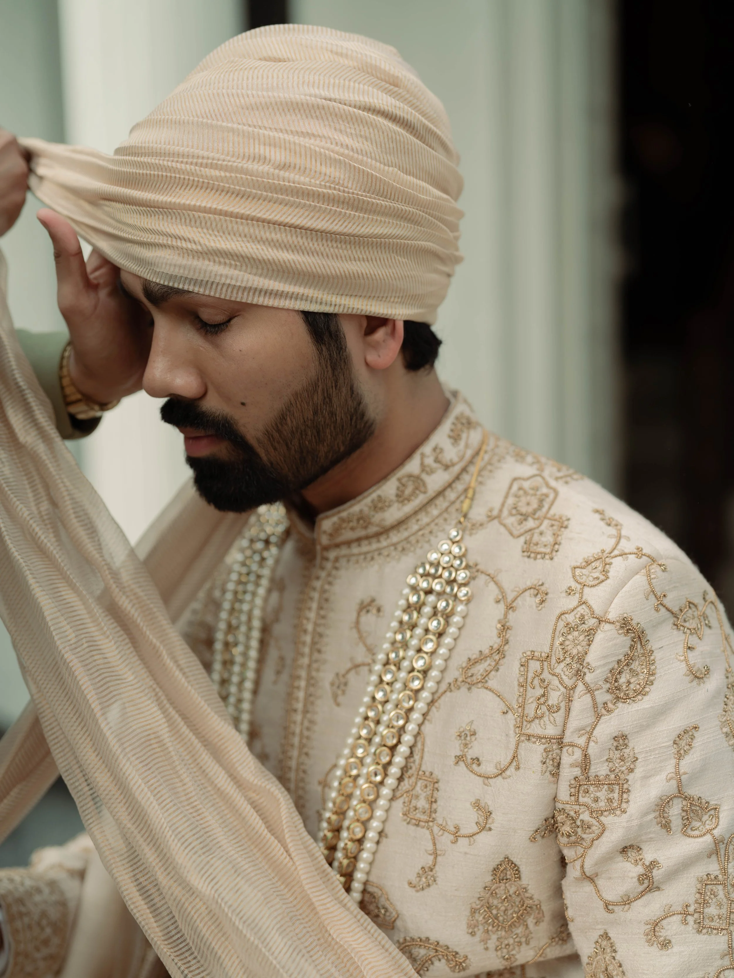 A man dressed in traditional Indian attire, wearing a beige turban, ornate cream-colored kurta with gold embroidery, and multiple pearl necklaces, standing with his eyes closed and hand resting on his forehead.