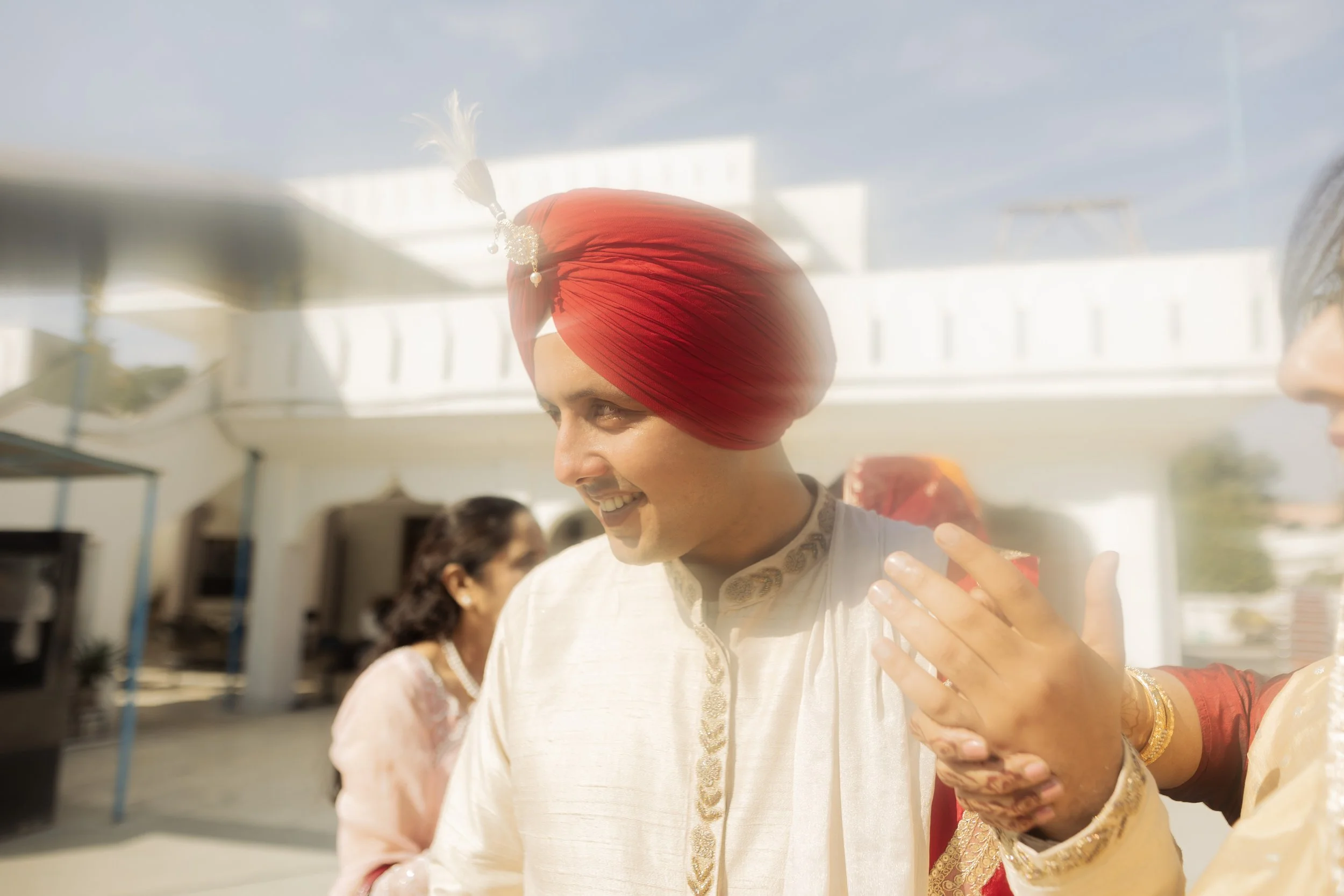 Man wearing a white traditional Indian outfit and red turban smiling at a welcoming event, with a woman in traditional attire greeting him and a woman in the background.