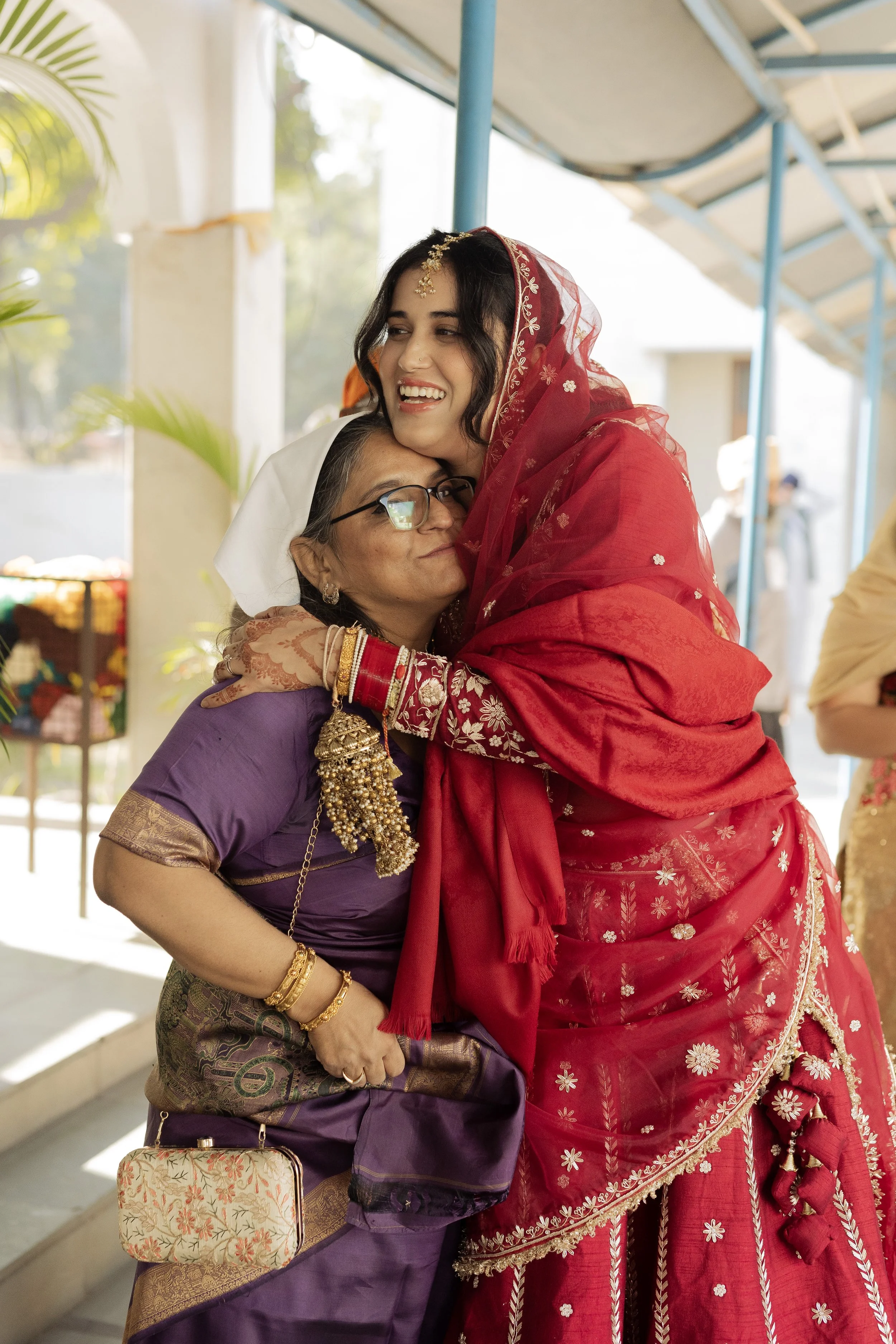 Two women embracing, one in traditional purple attire with jewelry and the other in a red embroidered saree and veil, at a festive occasion.