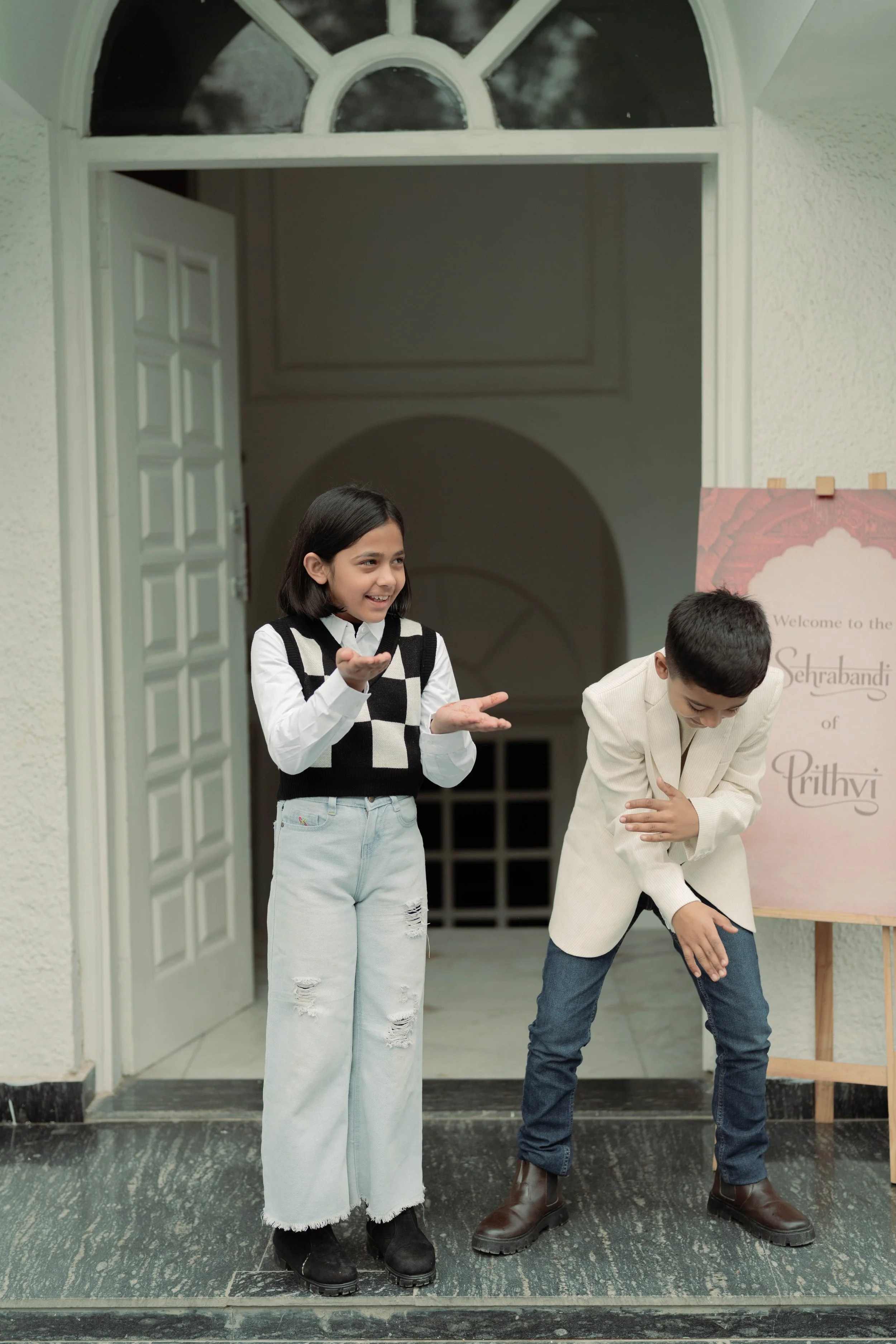 A girl and boy standing at the entrance of a house, laughing and talking. The girl has black hair, wearing a black and white checkered vest and jeans, while the boy is dressed in a cream-colored blazer and dark jeans, appearing to be enjoying a conve