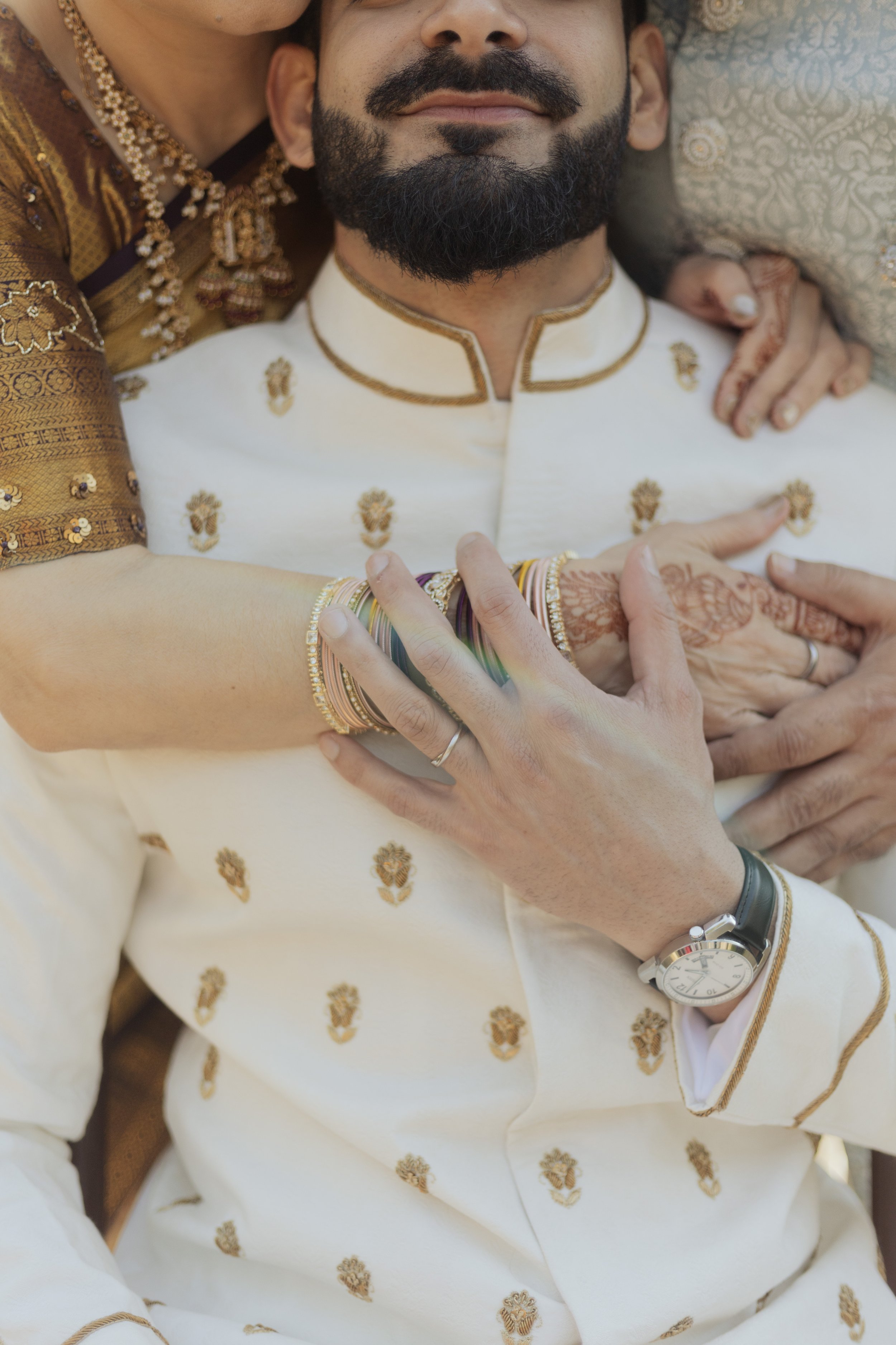 Close-up of a couple dressed in traditional Indian wedding attire, with the bride's hands and arms around the groom, showing intricate henna designs, bangles, rings, and jewelry.