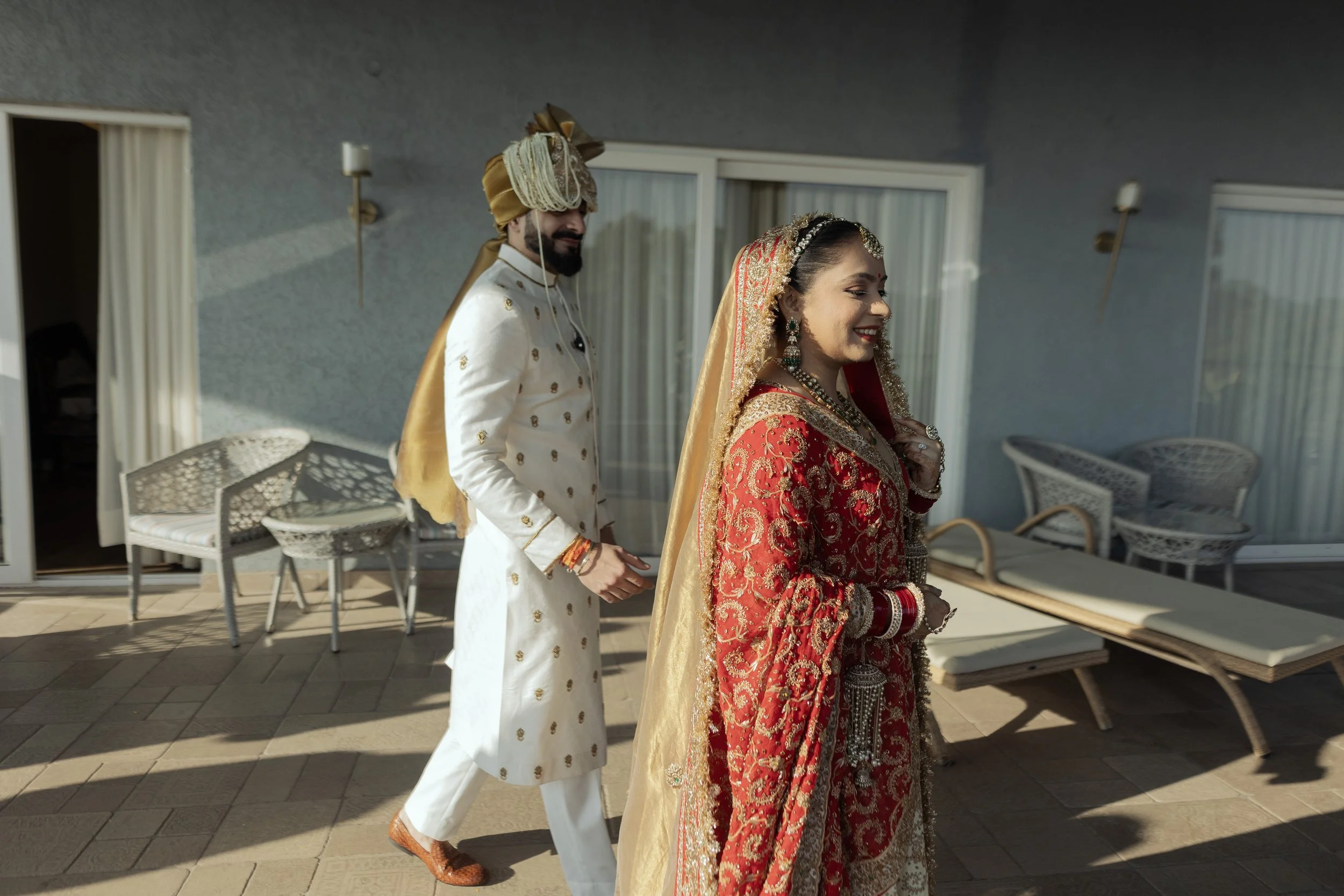 Indian bride in red traditional wedding attire and groom in cream and gold traditional attire standing outdoors on a patio.