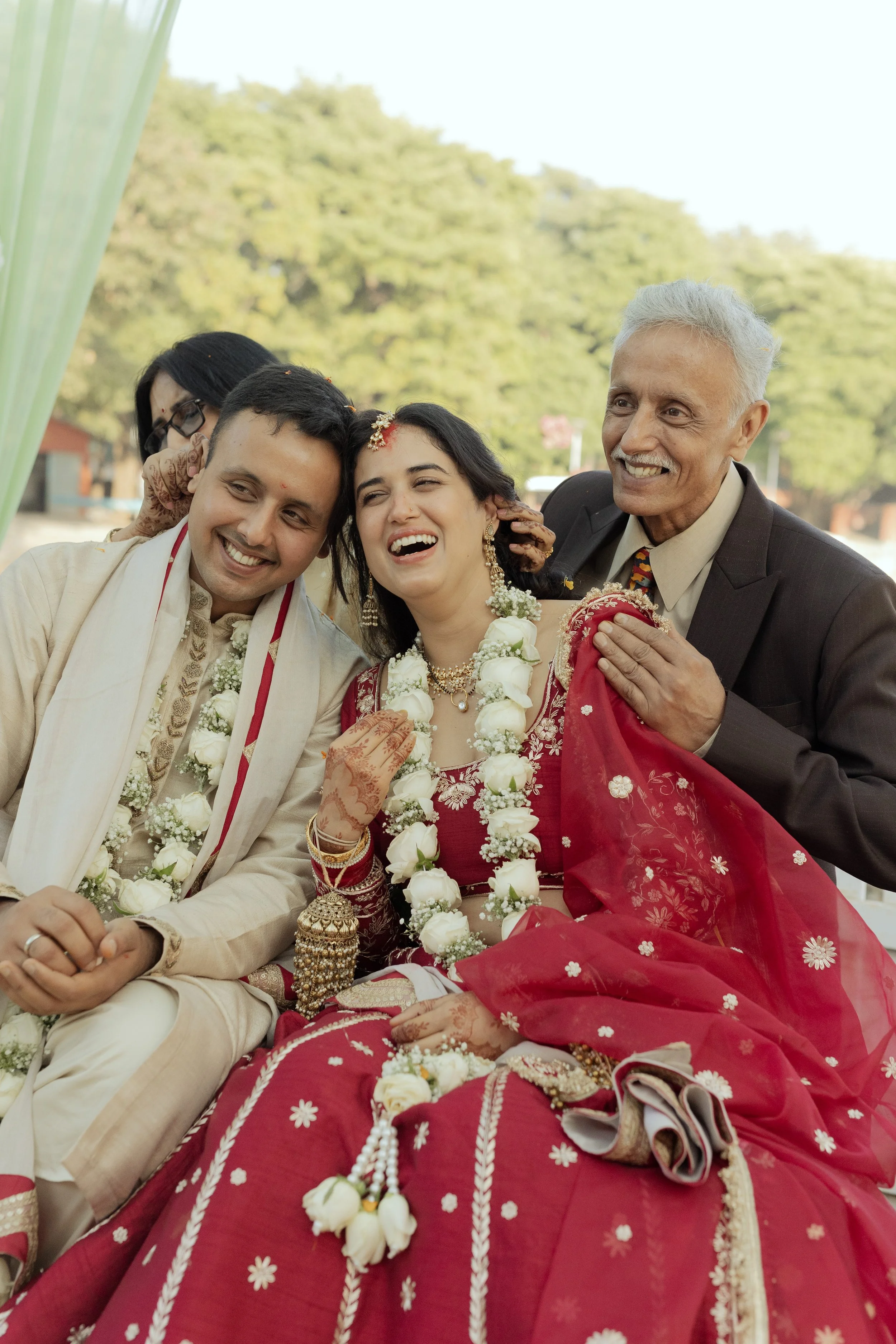 A happy Indian wedding celebration with a bride in a red saree and floral jewelry, flanked by a groom and an older man, all smiling and celebrating outdoors.