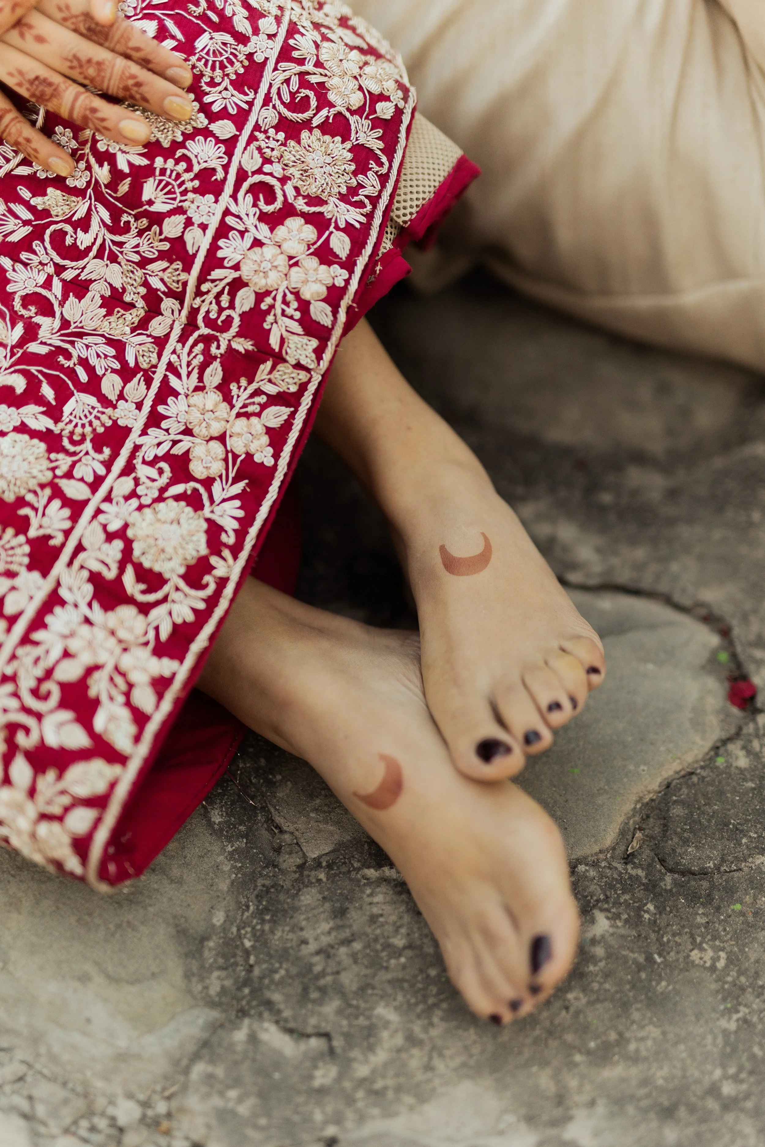 Close-up of a person's feet with henna tattoos, resting on a stone surface; they have dark painted toenails and are wearing traditional embroidered clothing with floral patterns.