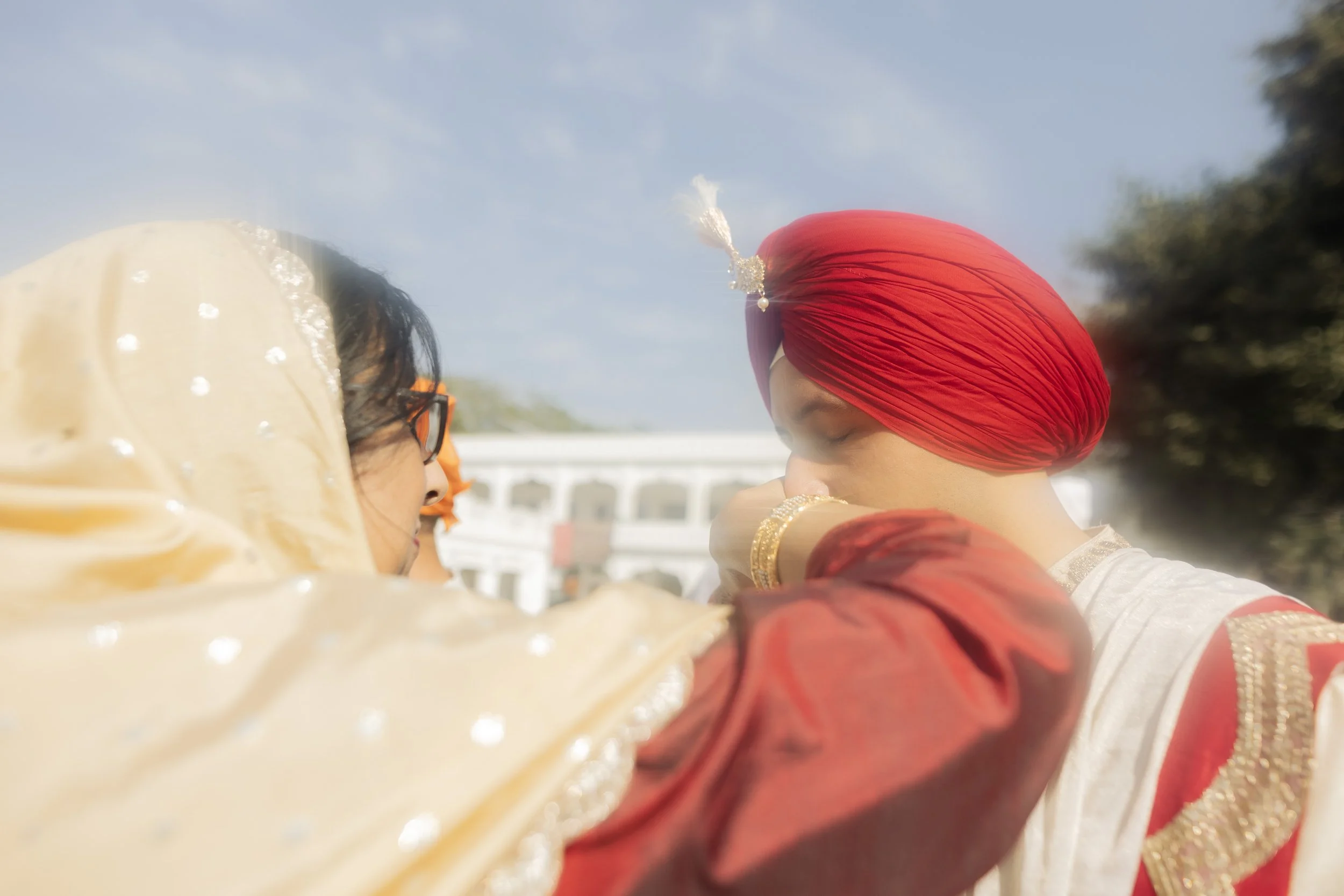 Two women, one wearing a traditional red turban and attire, and the other with a beige dupatta with white dots, are engaged in a ceremonial moment outdoors during daylight, with a building and trees in the background.