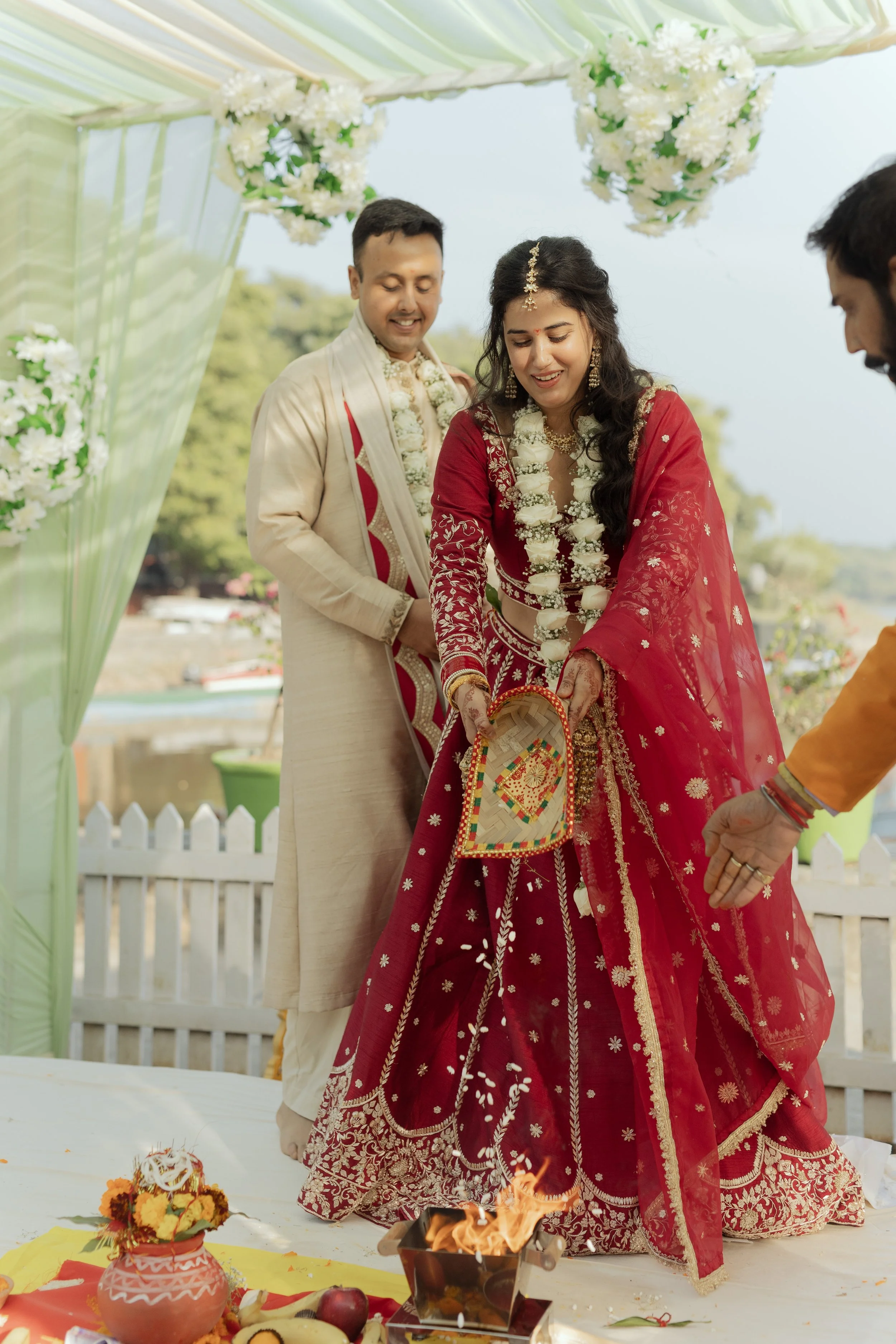 Indian wedding ceremony with a bride in a red saree, a groom, and two men, one of whom is lighting a sacred fire during the ritual, outdoors decorated with flowers and a white picket fence.