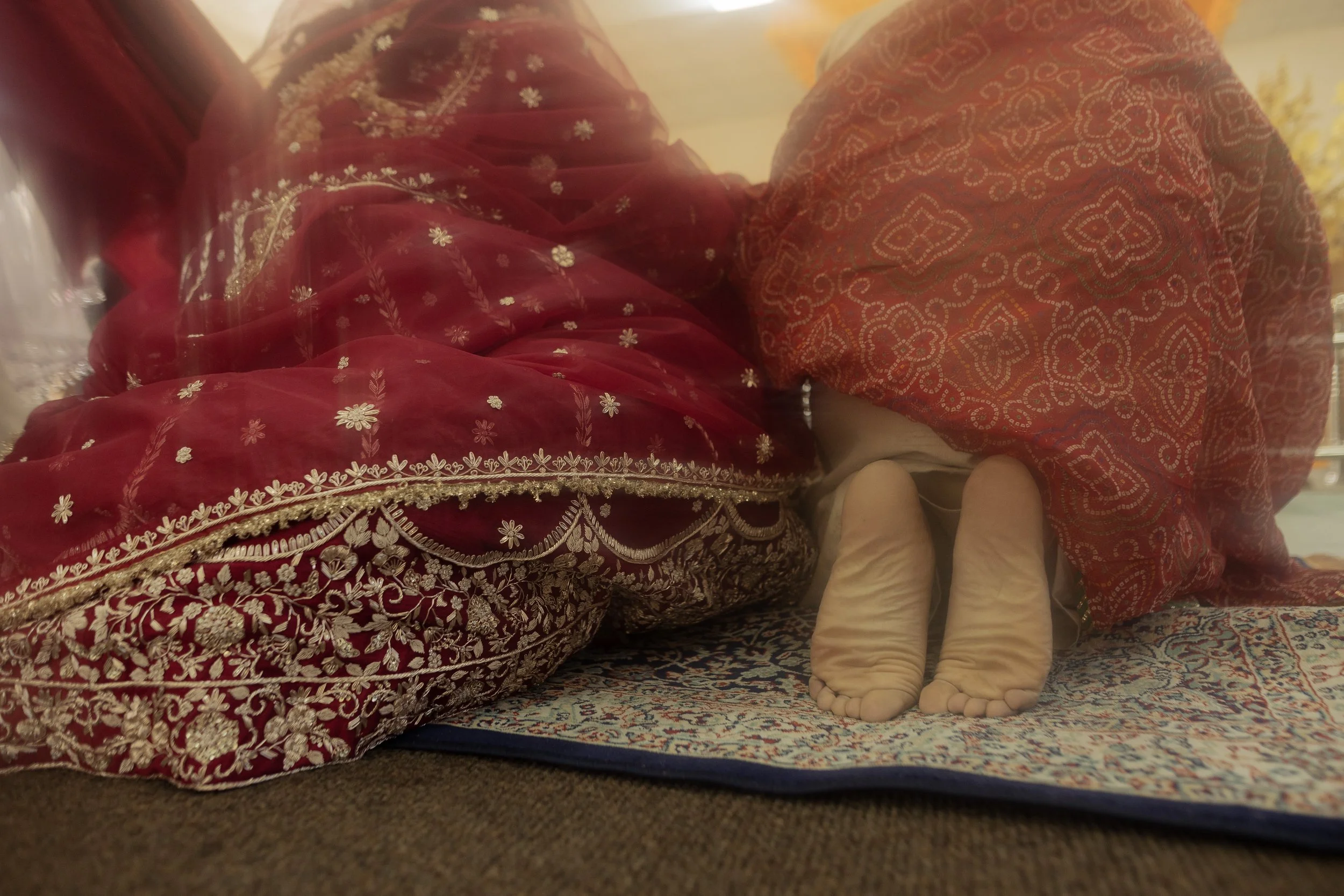 Child dressed in traditional Indian attire with a detailed embroidered red and gold outfit, hiding under a matching red scarf, standing on a patterned rug.
